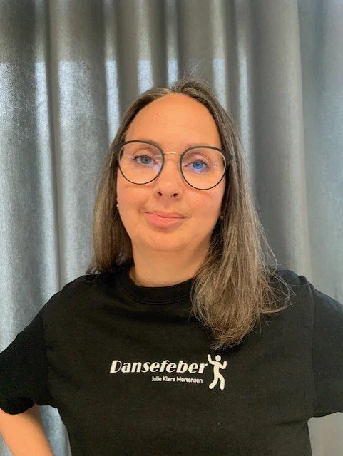 Kvinde med briller, iført sort t-shirt med teksten 'Dansefeber' og en gestus med armene ud til siderne, foran en grå gardinbaggrund. Danseskole i Aarhus