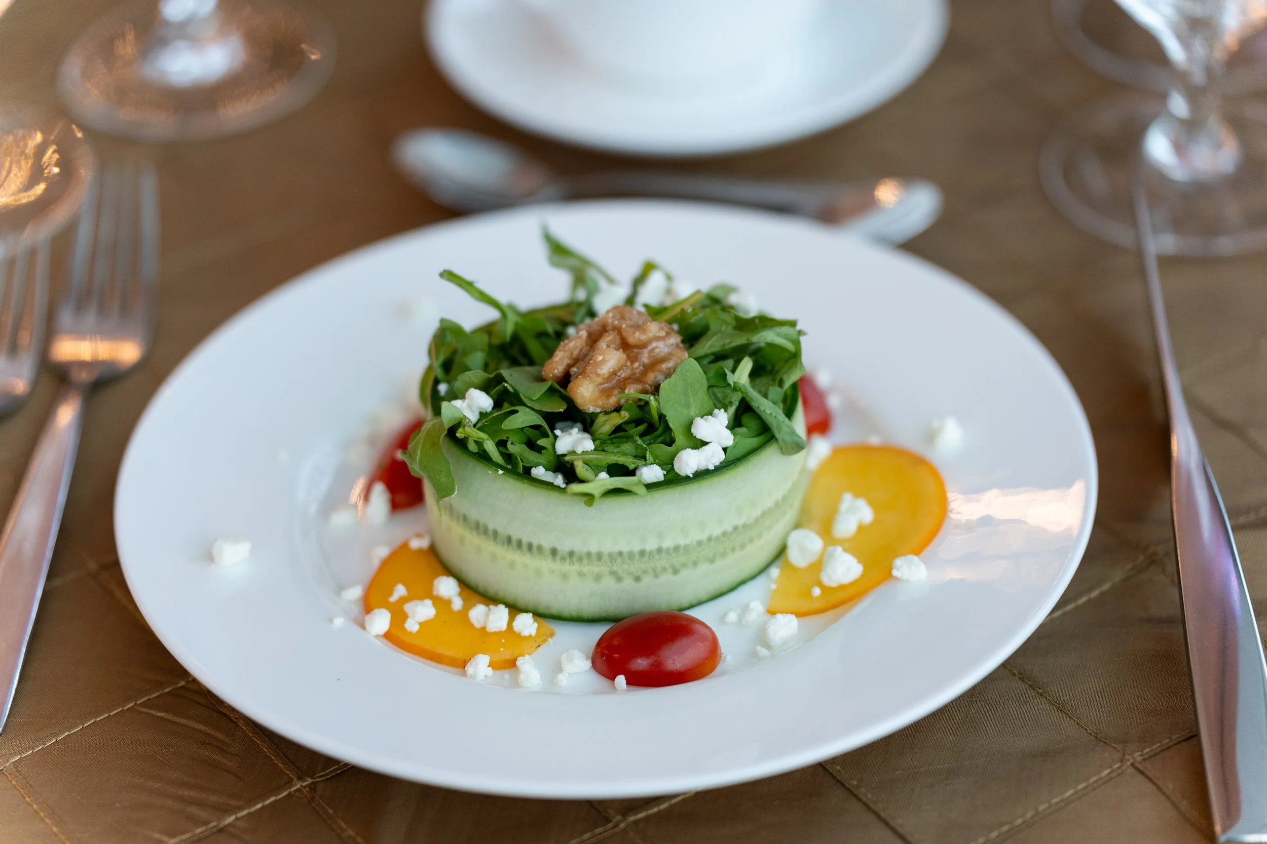 A fresh vegetable salad with sliced cucumber, cherry tomatoes, greens, walnuts, crumbled cheese, and yellow bell pepper slices on a white plate.