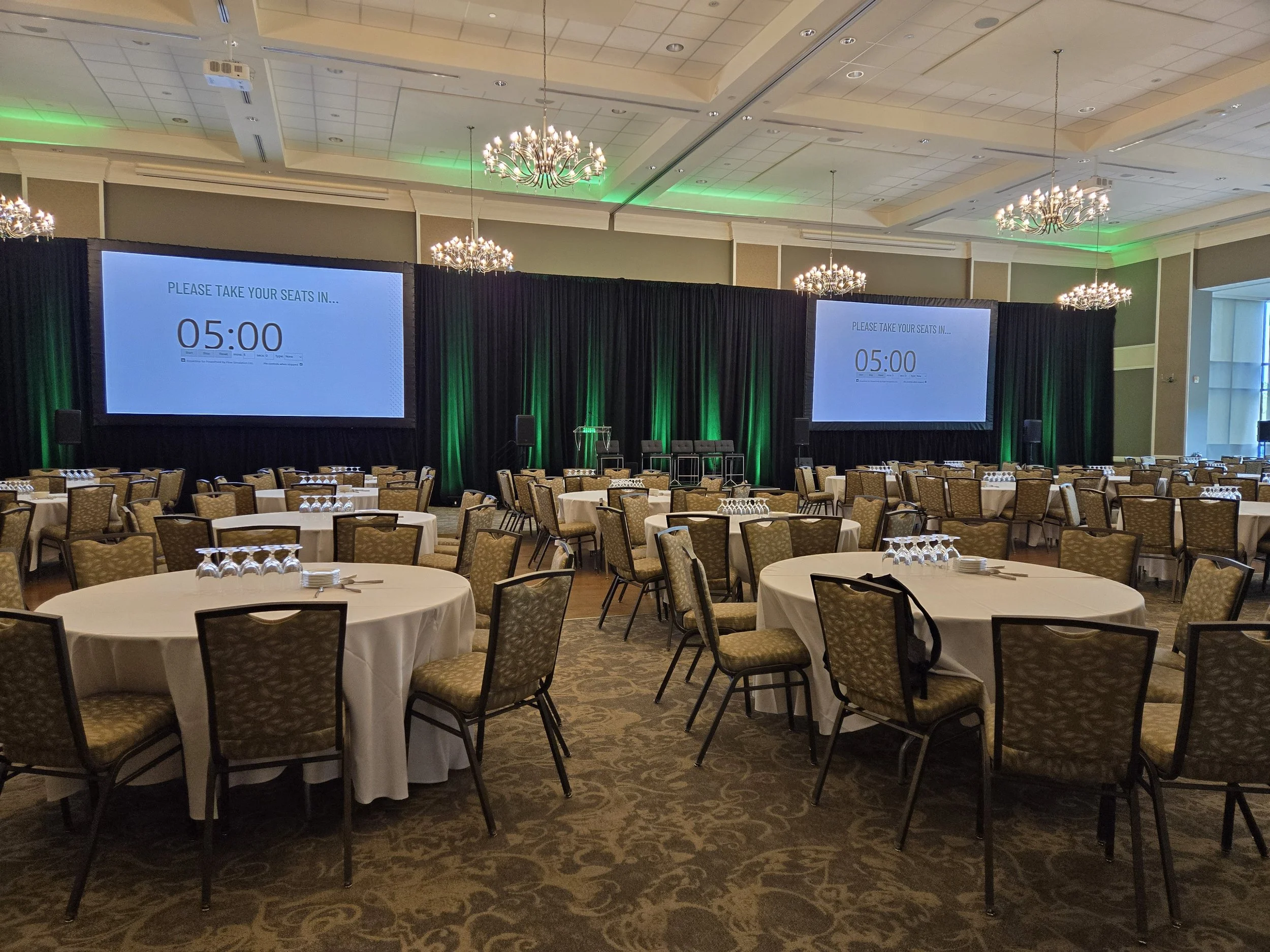 Conference room with round tables set for an event, two large screens display a countdown timer and a message for attendees, chandeliers hang from the ceiling, and stage with black curtains and stanchions is at the front.
