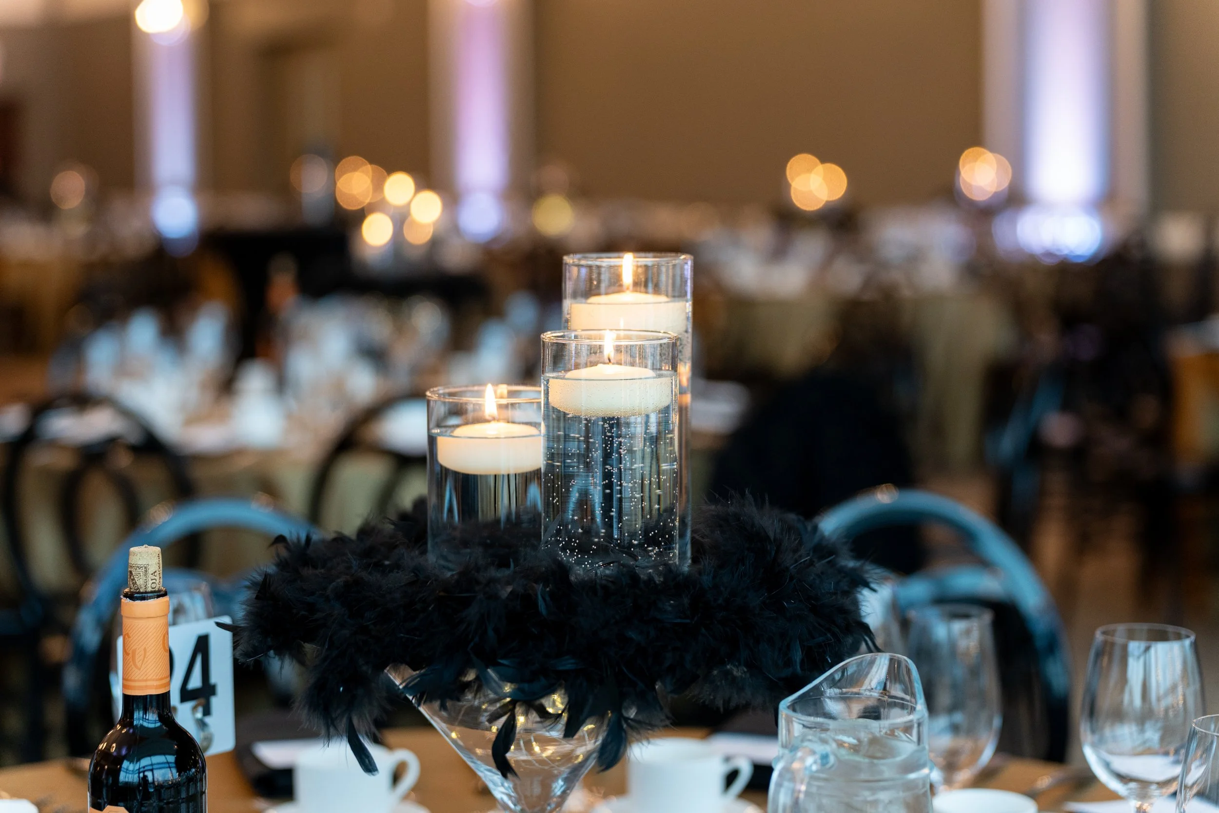 Centerpiece with three lit candles in glass vases on a table at an event, with a blurred background of other tables and warm lighting.