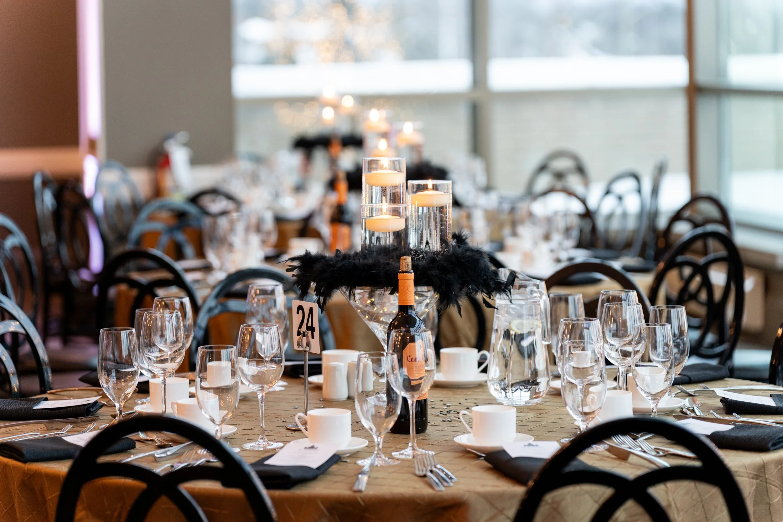 A decorated dining table for a formal event with candles, wine bottles, glassware, and black napkins, set in a room with large windows and chairs around the table.