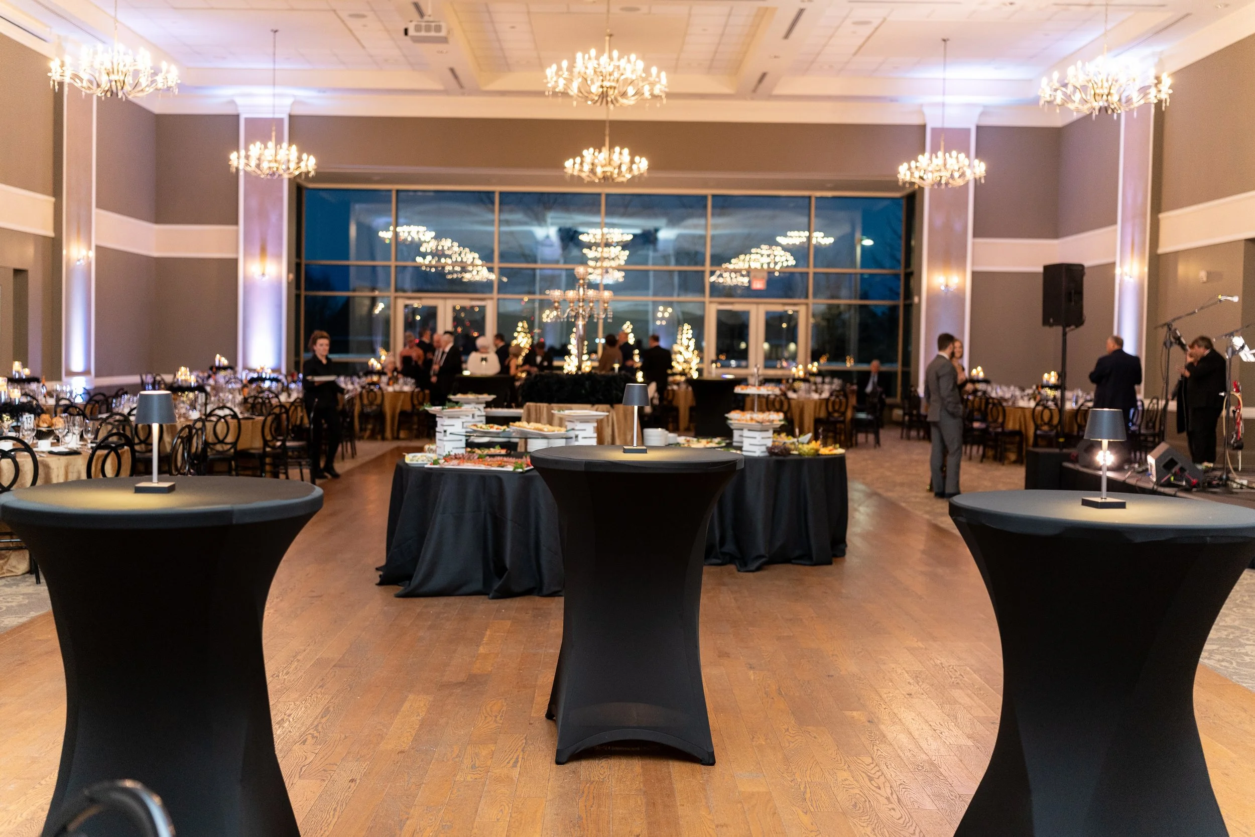 A banquet hall decorated for a formal event with round tables, black tablecloths, and elegant chandeliers hanging from the ceiling. The hall features large windows reflecting more chandeliers, and a buffet table with food and supplies is set up in th