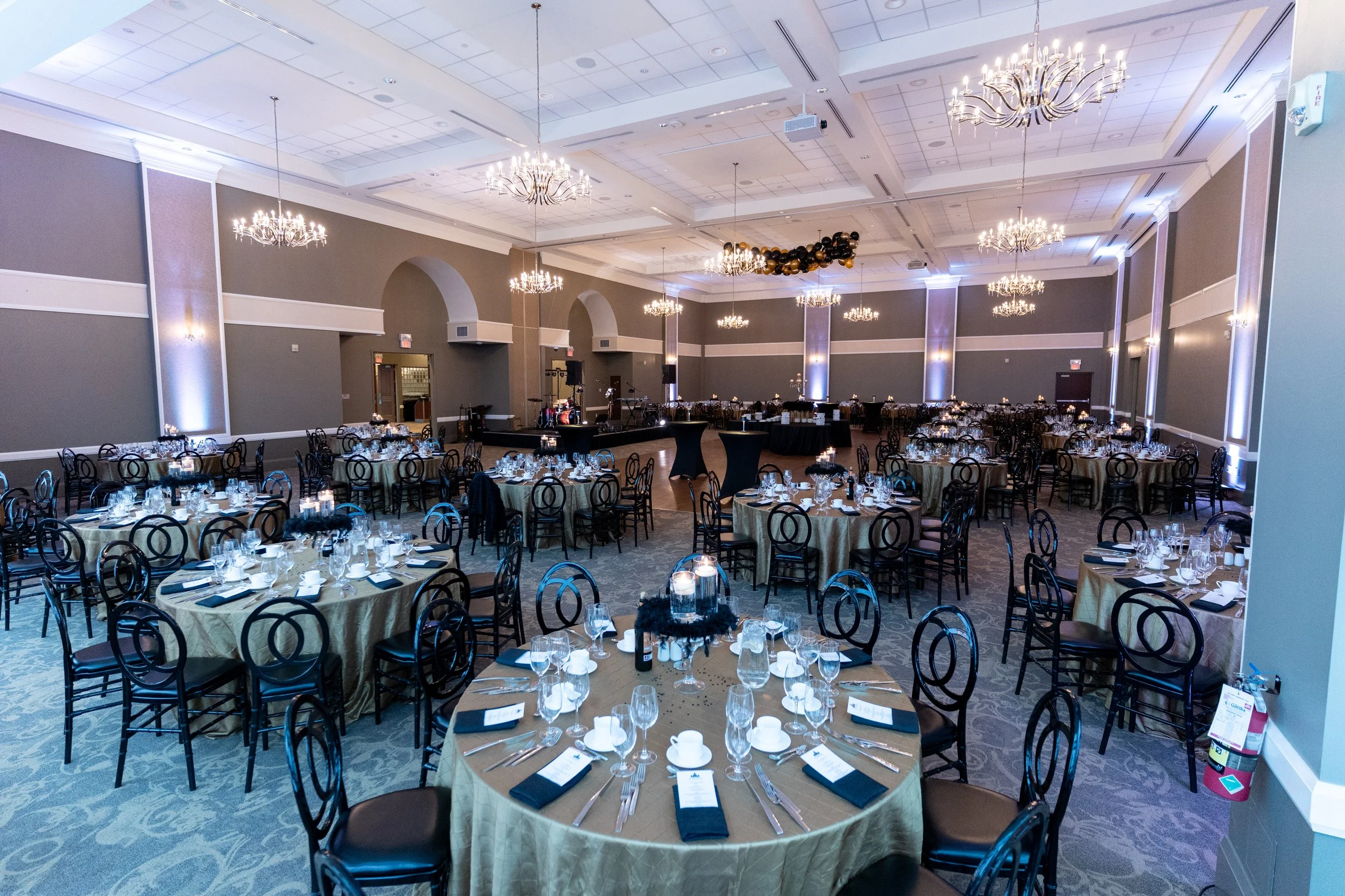 Elegant banquet hall decorated for a formal event with round tables set with beige tablecloths, black chairs, glassware, and napkins, chandeliers hanging from the ceiling, stage with musical instruments, dark balloons and gold decorations, and soft l