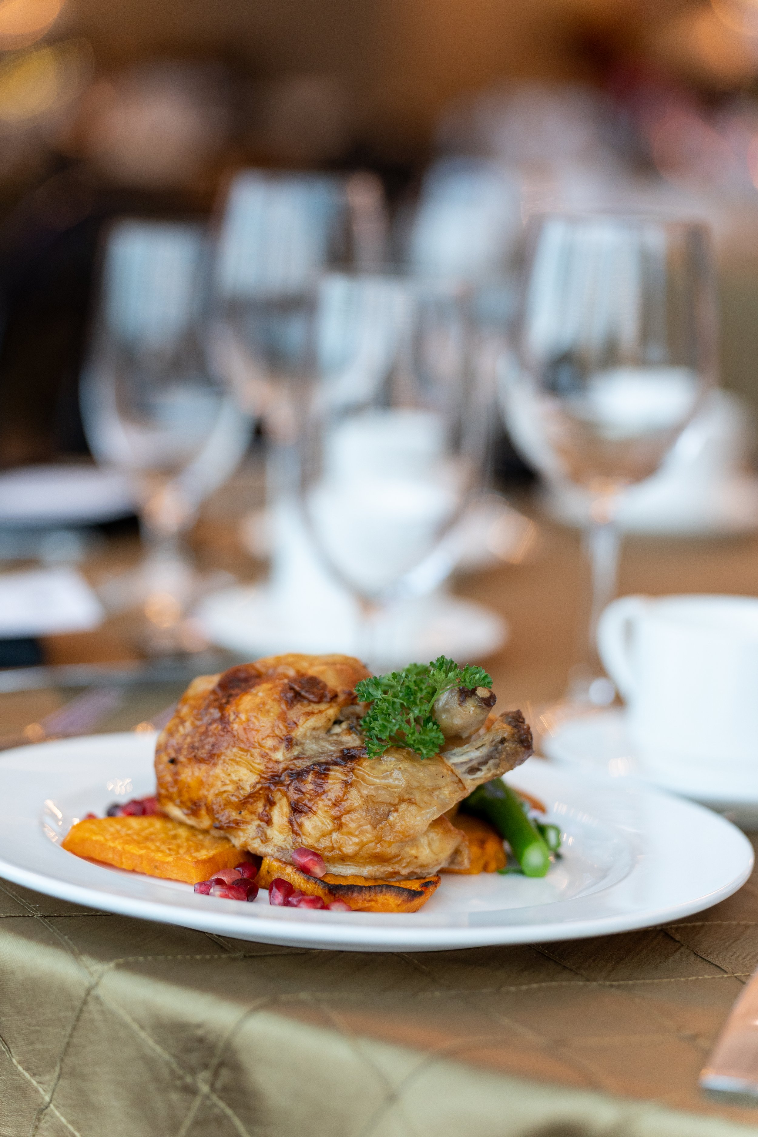 Close-up of a plated roasted chicken leg garnished with parsley, served with roasted vegetables, with empty wine glasses in the background.