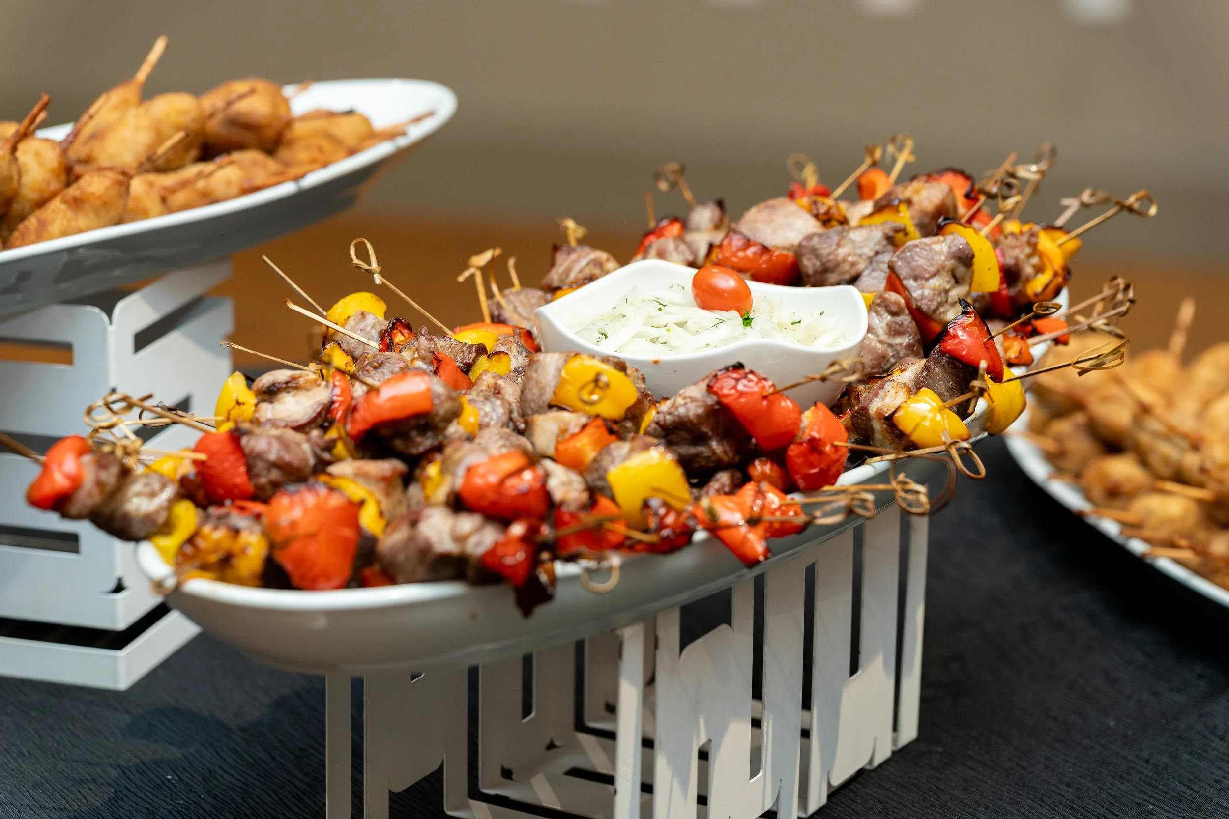 A white serving tray with skewers of grilled meat and colorful peppers, accompanied by a small bowl of coleslaw, on a black table.