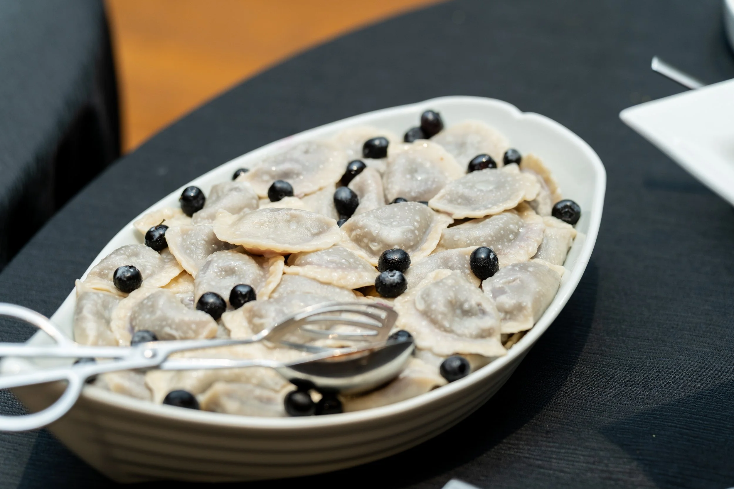 A white oval dish filled with boiled dumplings topped with black berries on a dark table.