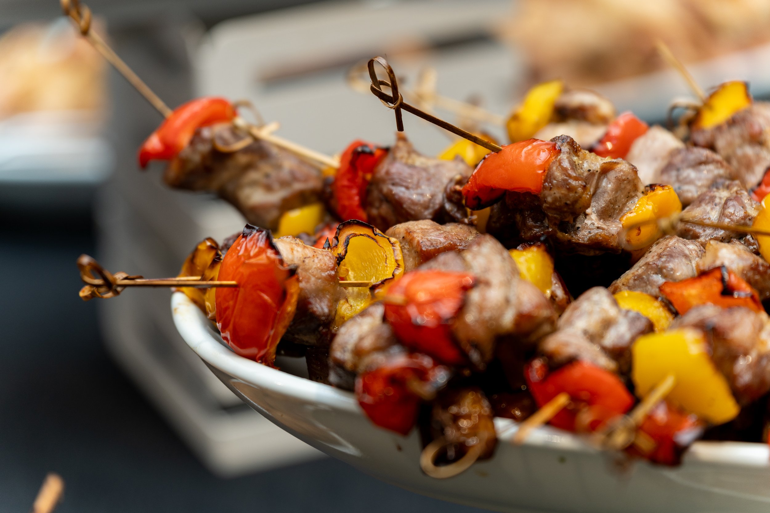 Close-up of skewers with grilled chunks of meat, red and yellow bell peppers, and cherry tomatoes in a white bowl.