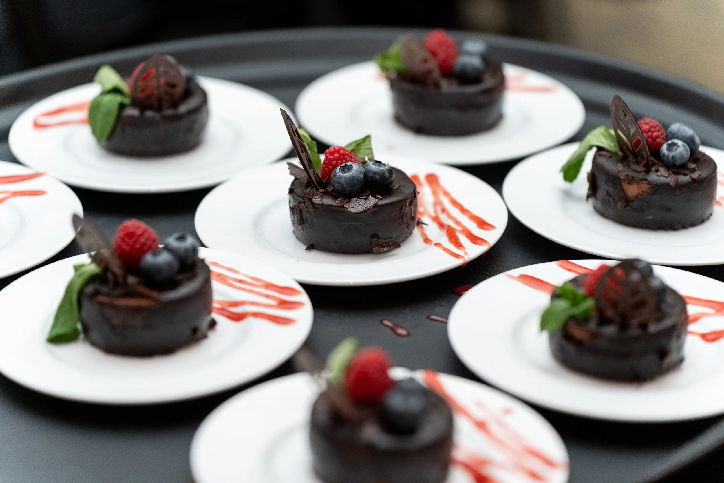 A tray of individual chocolate desserts topped with blueberries, raspberries, and a green mint leaf, garnished with a chocolate piece and drizzled with red sauce.