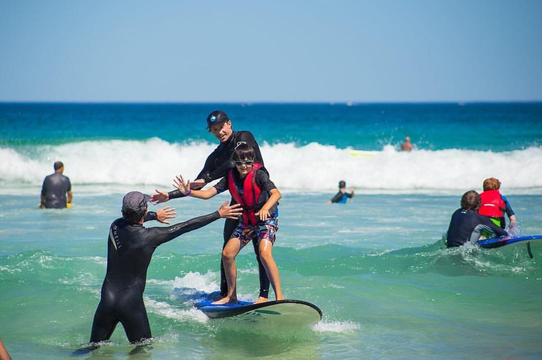 Surf instructors helping a young beginner stand up on a surfboard during a kids' surf lesson in Perth, Western Australia