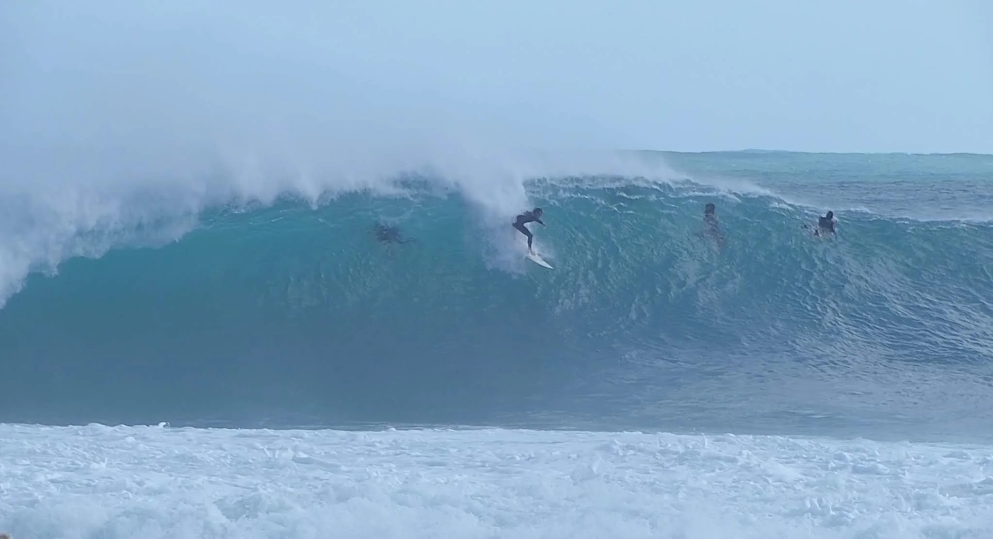 Surfer riding a large wave at a powerful reef break in Western Australia, with other surfers paddling in the lineup