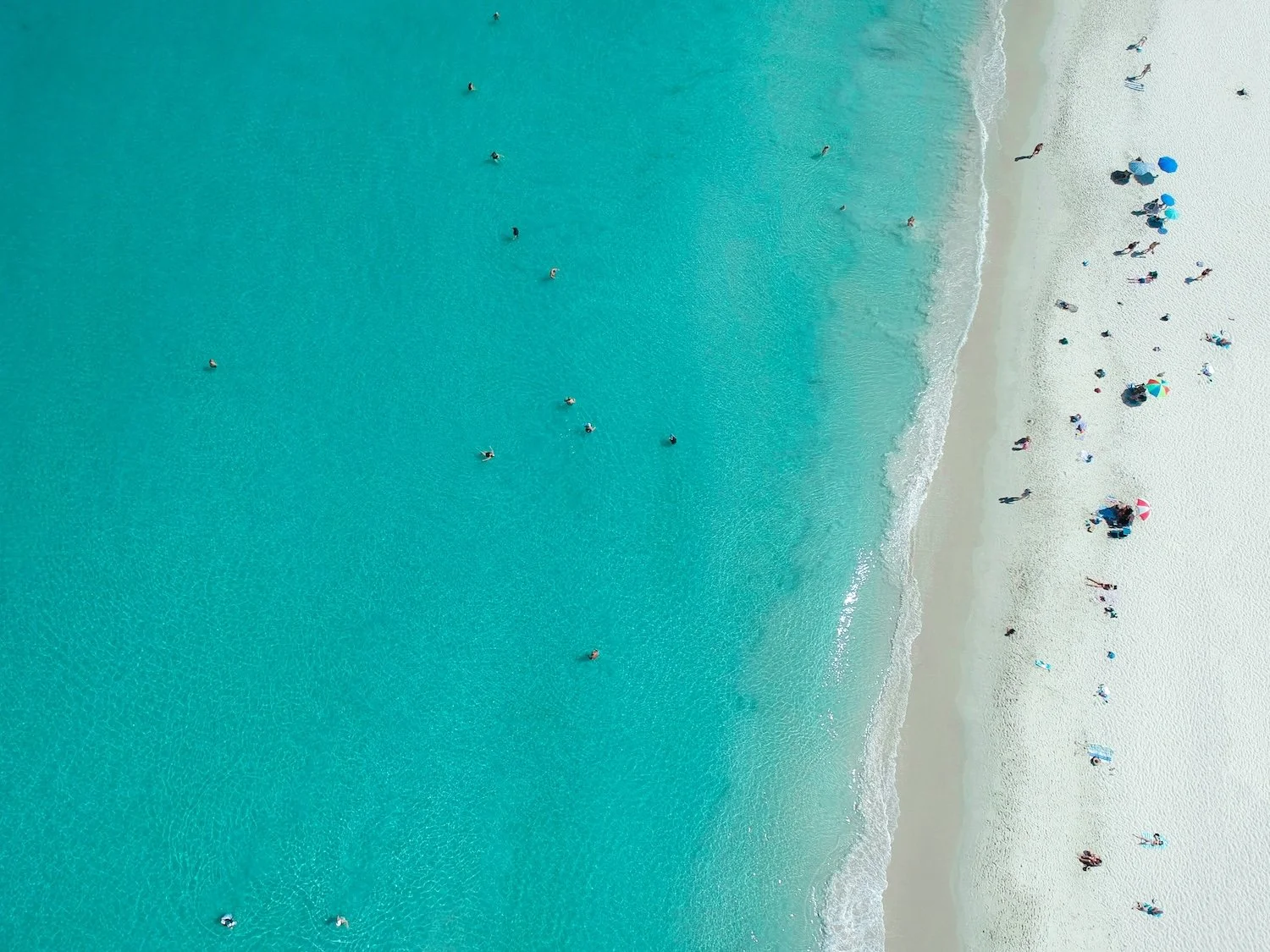 Aerial view of a crowded beach with people sunbathing, swimming, and walking along the shoreline.