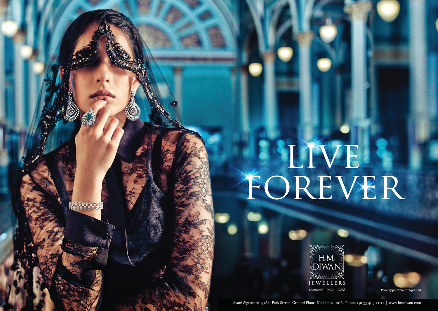 A woman dressed in black lace clothing and jewelry, covering her eyes with a lace veil, stands in an ornate, brightly lit indoor setting with an arched ceiling. The text reads "Live Forever" and an advertisement for H.M. Diwan Jewellers.