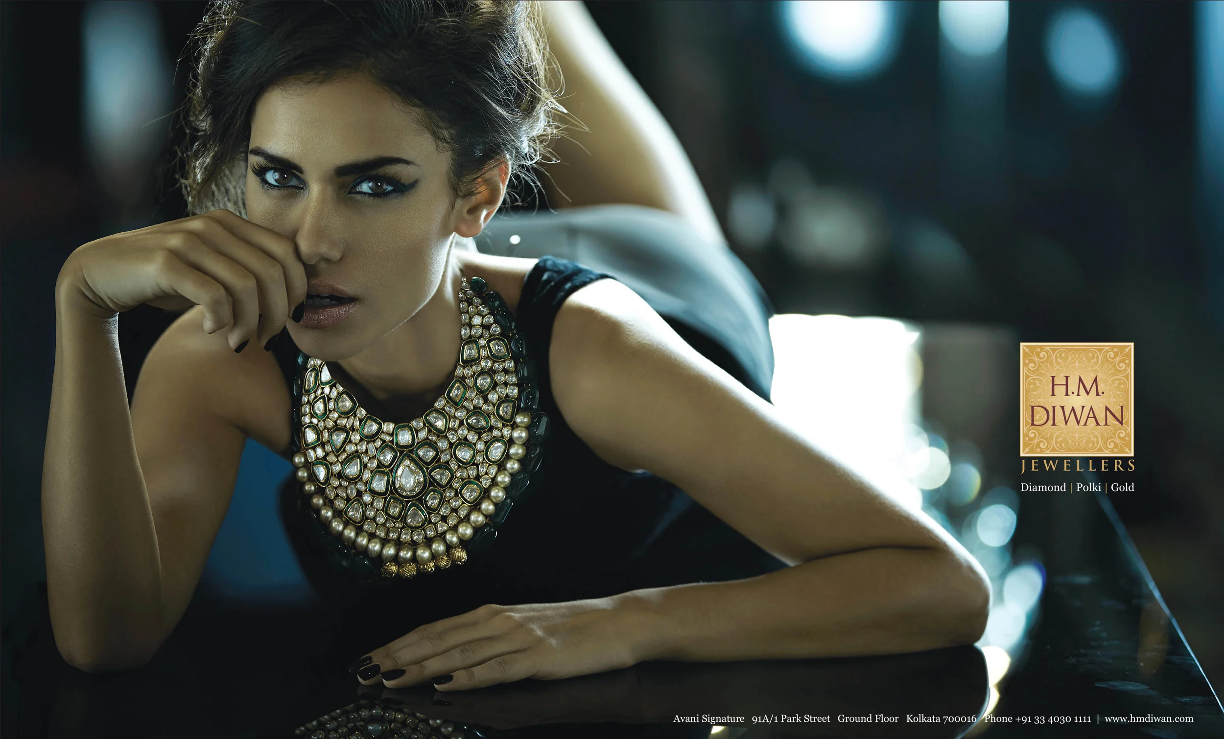 A woman with dark hair and striking blue eyes wearing a large, elaborate diamond and pearl necklace, lying on a dark reflective surface with a blurred bokeh background, promoting H.M. Diwan Jewellers.