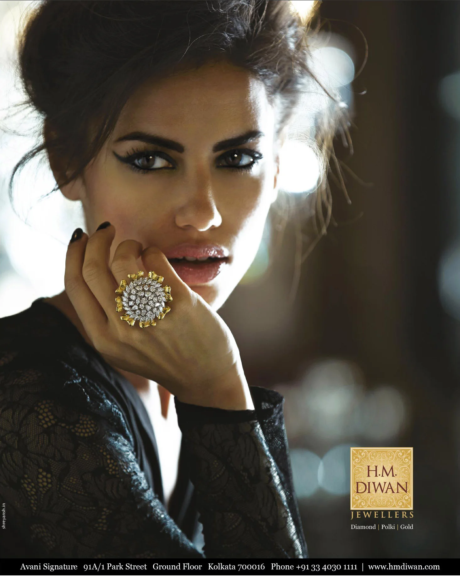 A woman with dark hair, smoky eye makeup, and black lace clothing, wearing a large gold and diamond ring, holding her hand near her face, with a blurred background and a H.M. Diwan Jewellers logo at the bottom right corner.