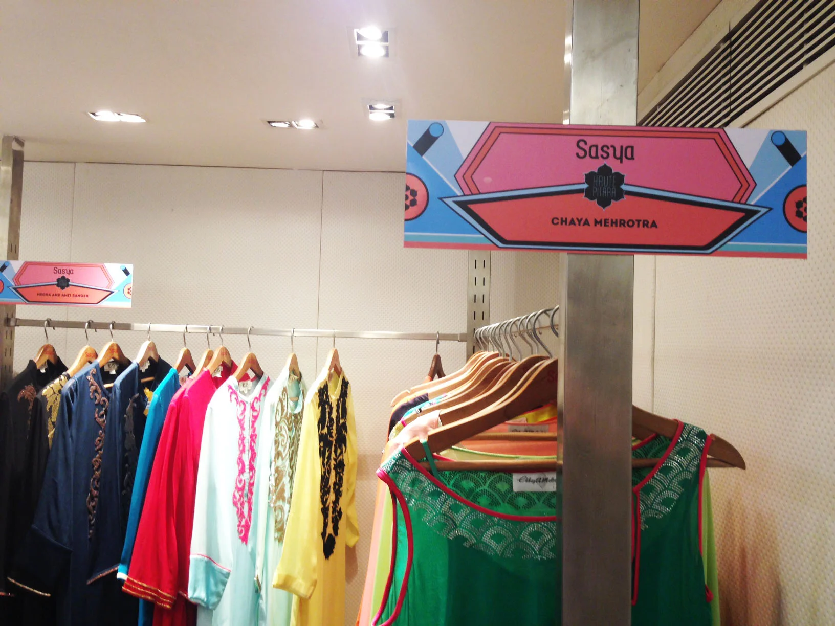 Clothing rack with various colorful garments and a sign reading "Sasya" and "Chaya Mehrotra" in a store or boutique.