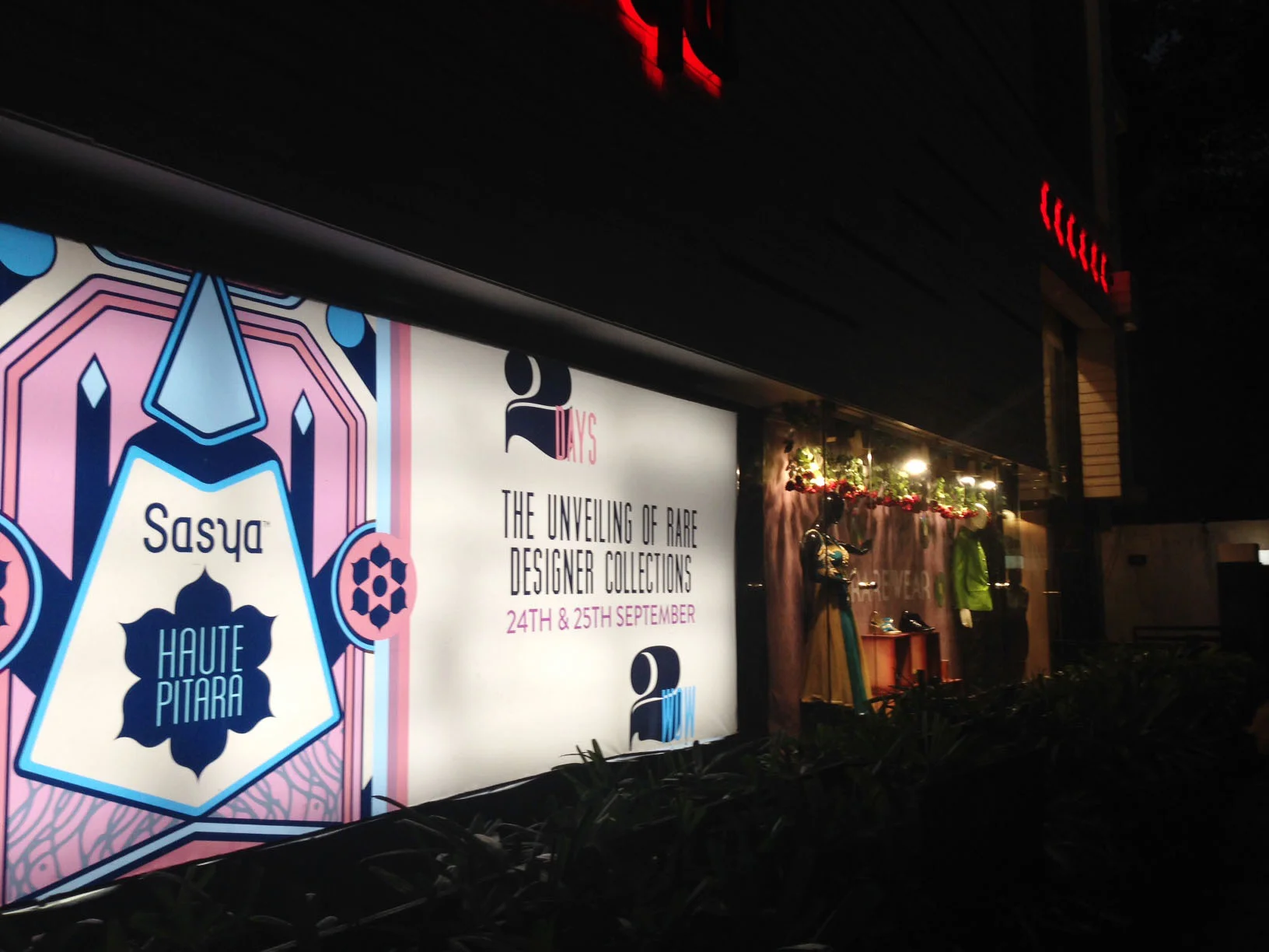 A large billboard advertisement for a designer collection event happening on September 24th and 25th. The billboard features colorful, geometric artwork and text promoting a fashion unveiling. To the right, there is a store window display with manneq