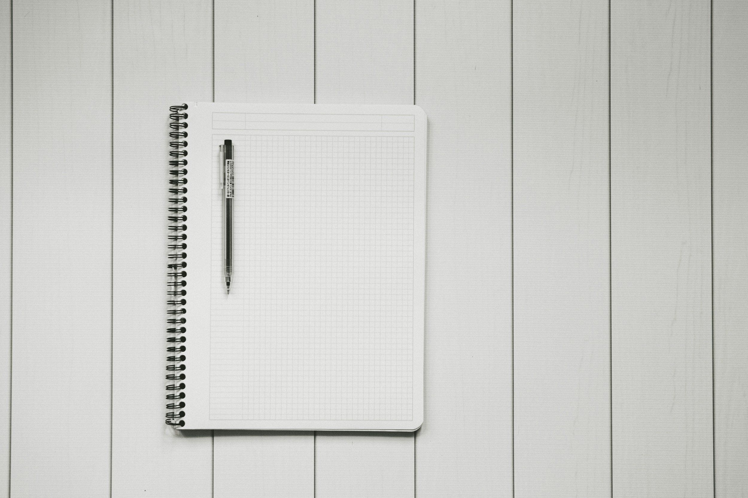 Open notebook with blank graph paper pages, a black and white pen, and a spiral binding, placed on a white wooden surface.