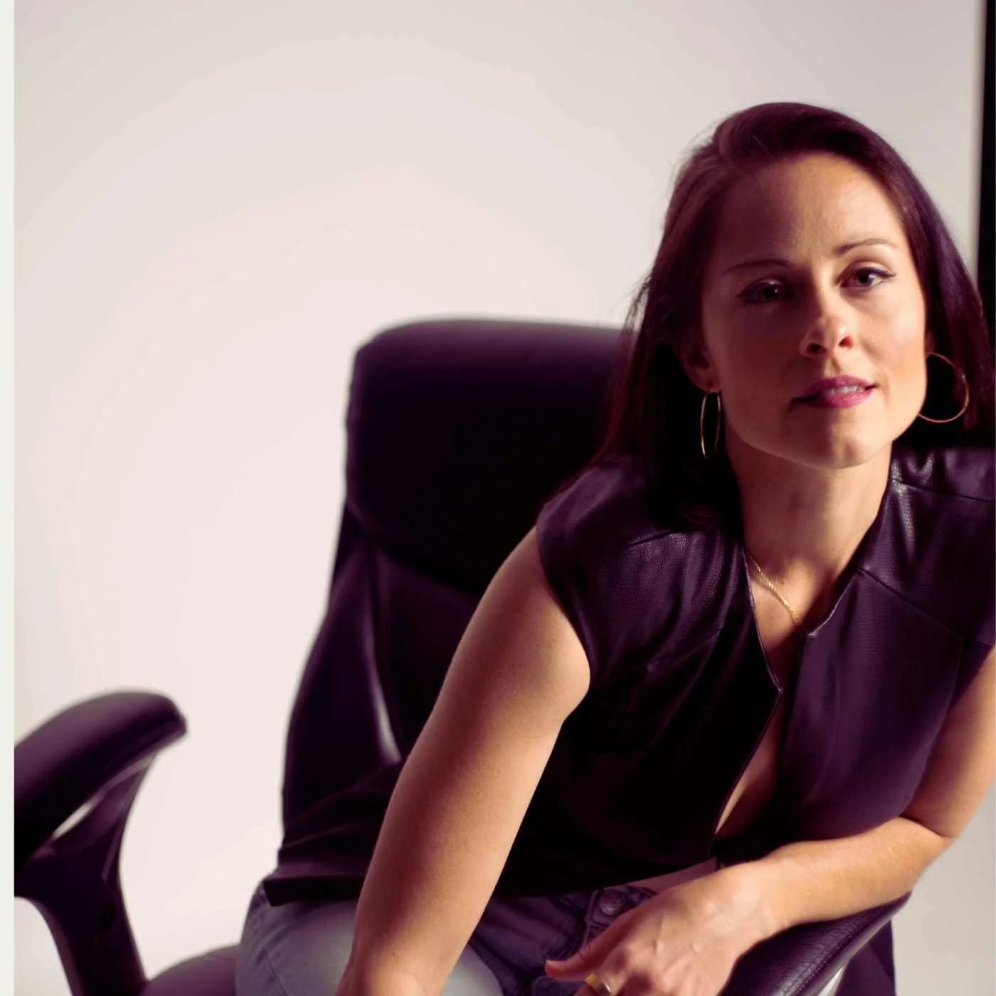 A woman with dark brown hair and hoop earrings sitting on a black office chair wearing a dark purple sleeveless top.