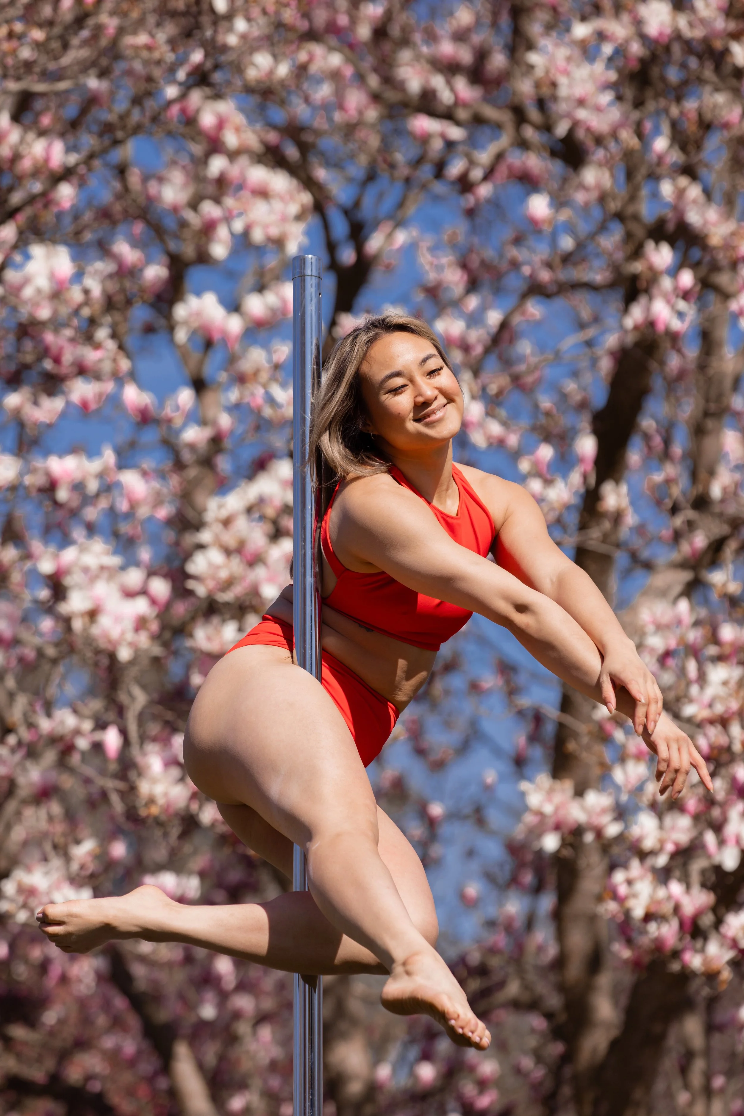Pole dancer performing outdoors in front of blooming magnolia trees during a spring photoshoot.