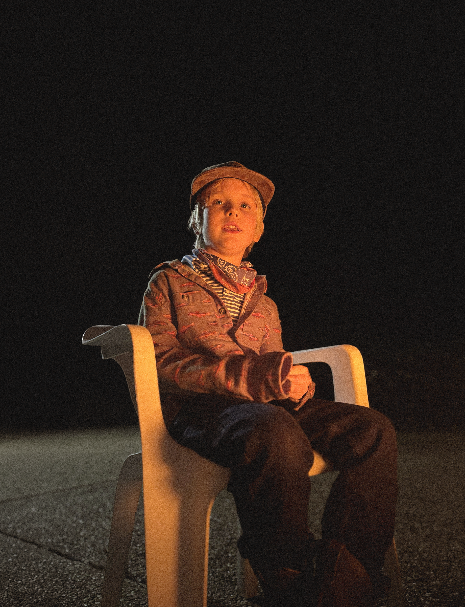 A young child with red hair sitting on a yellow plastic chair outdoors at night, illuminated by a nearby fire. The child is wearing a patterned jacket, a striped shirt, a scarf, and a cap, looking slightly upwards.
