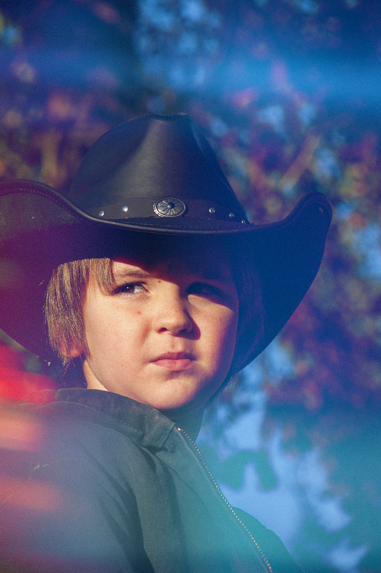 Close-up of a young person wearing a black cowboy hat and black jacket, looking into the distance with a serious expression, with a colorful and blurred background of trees and sky.