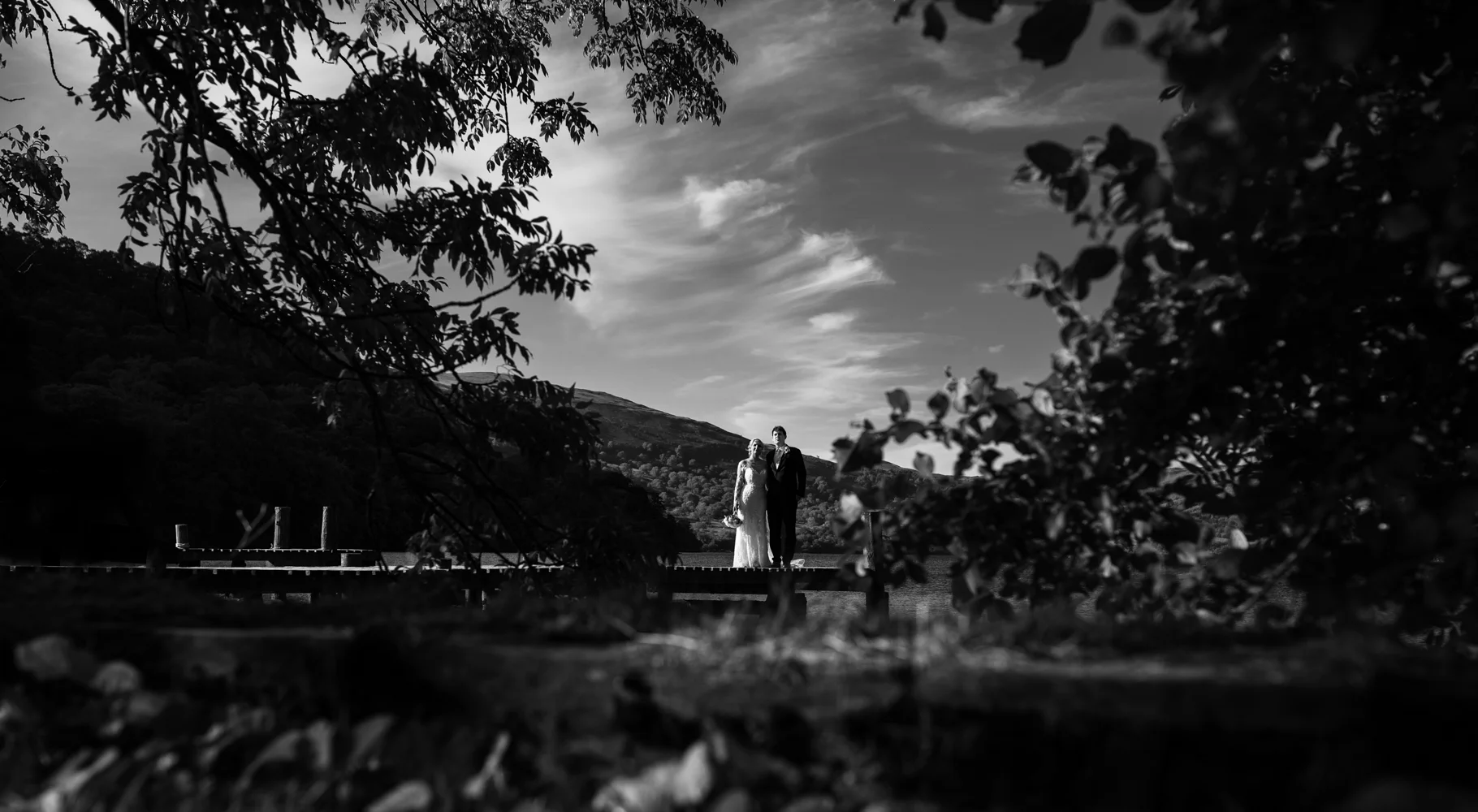 Black and white image of the bride and groom standing on the jetty taken through the trees at the waters edge