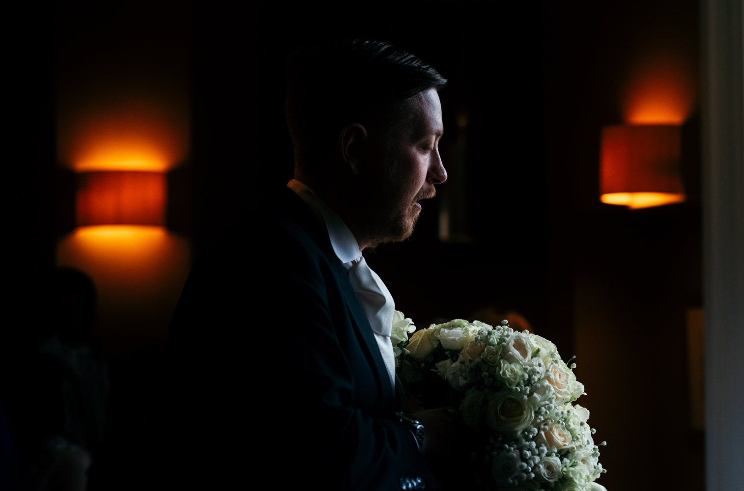 A dark and moody photo of the groom inside the wedding venue holding the flowers