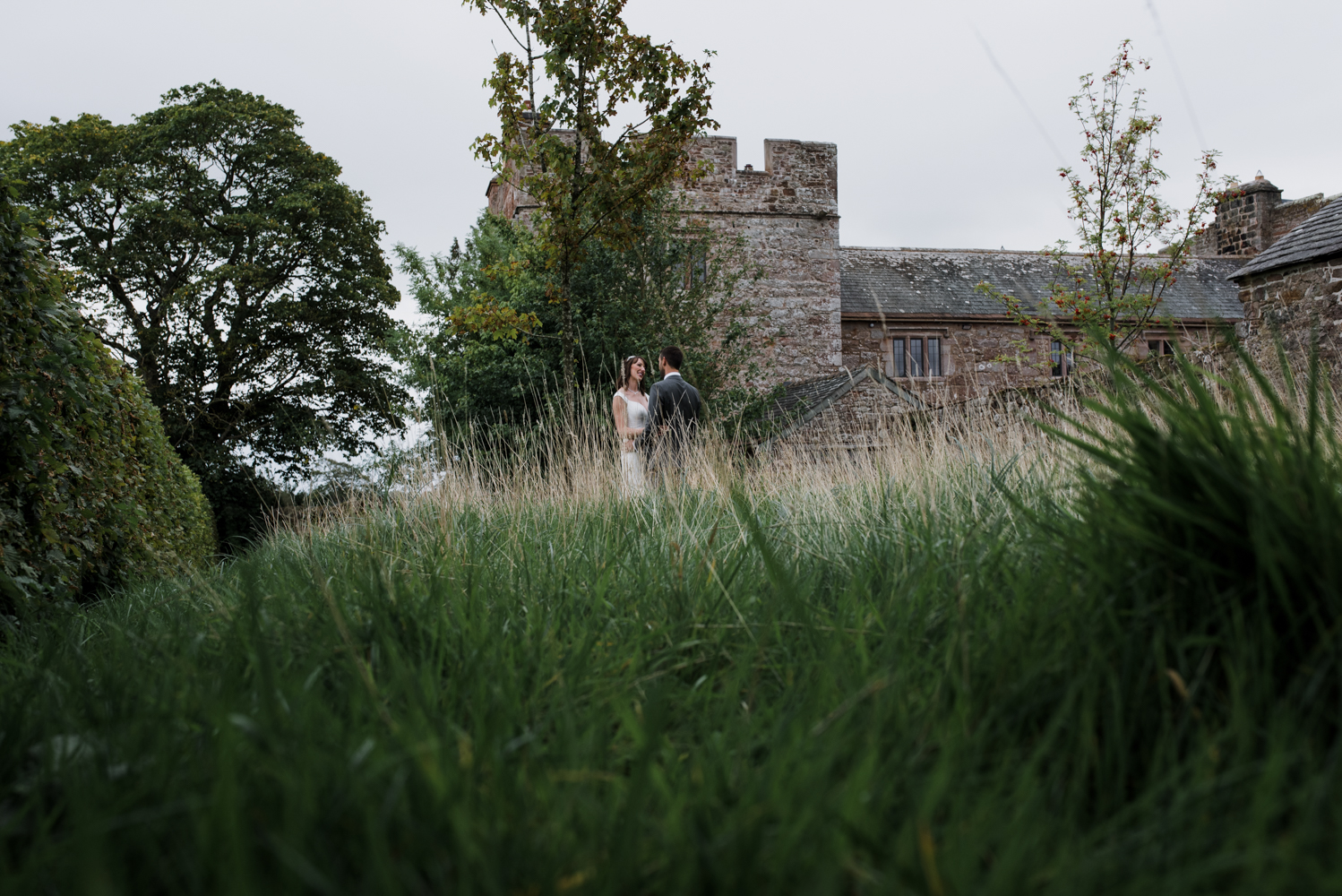 The bride and groom standing in the meadow with Blencowe Hall in the background