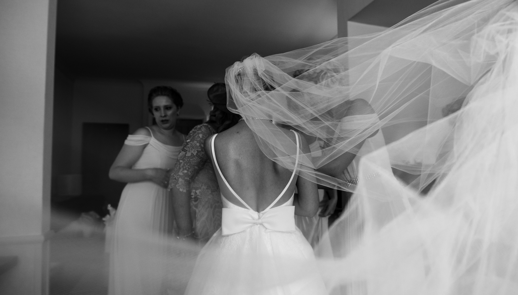 A black and white photo of the back of the brides dress with the veil flowing behind her