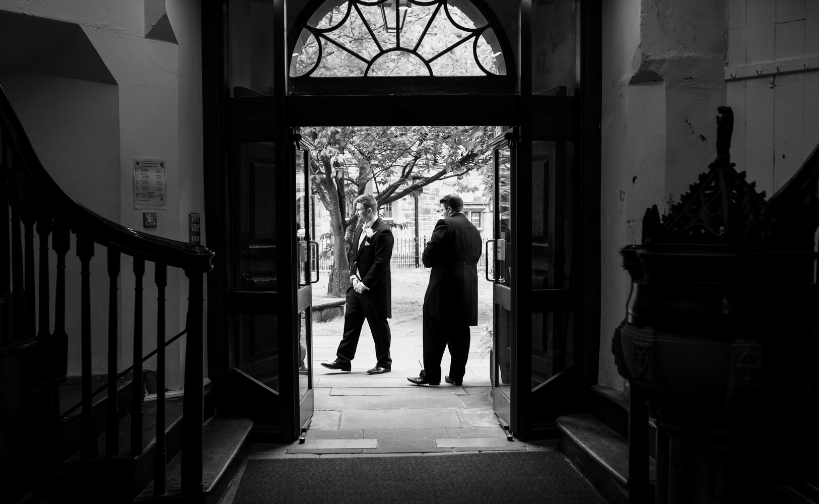 A black and white photo of the groom and his best man welcoming family and guests into the church before the ceremony