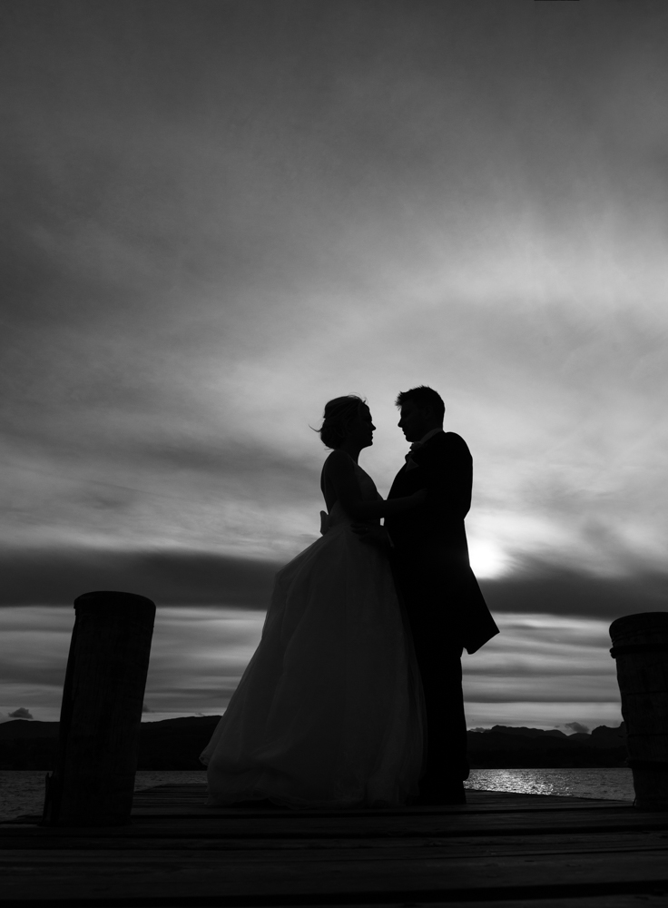 A black and white silhouette photo of the bride and groom standing on the jetty on the shoreline 