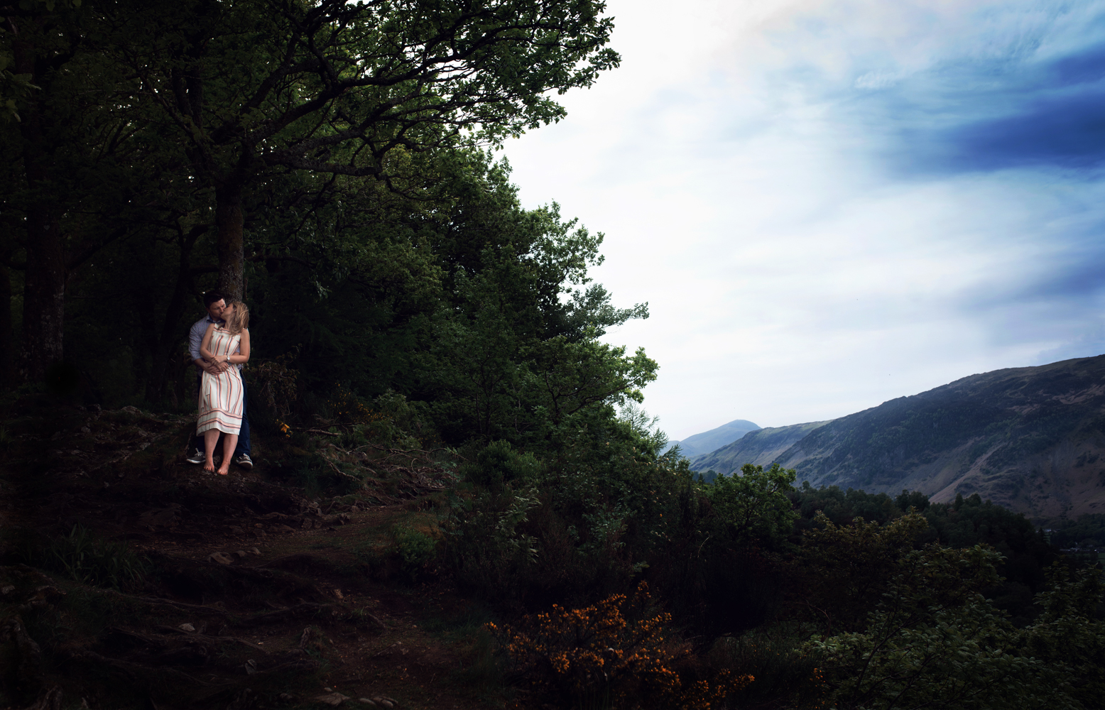 Pre shoot - A wide angle distant photo of a couple standing in the trees overlooking Derwent Water in the Lake District