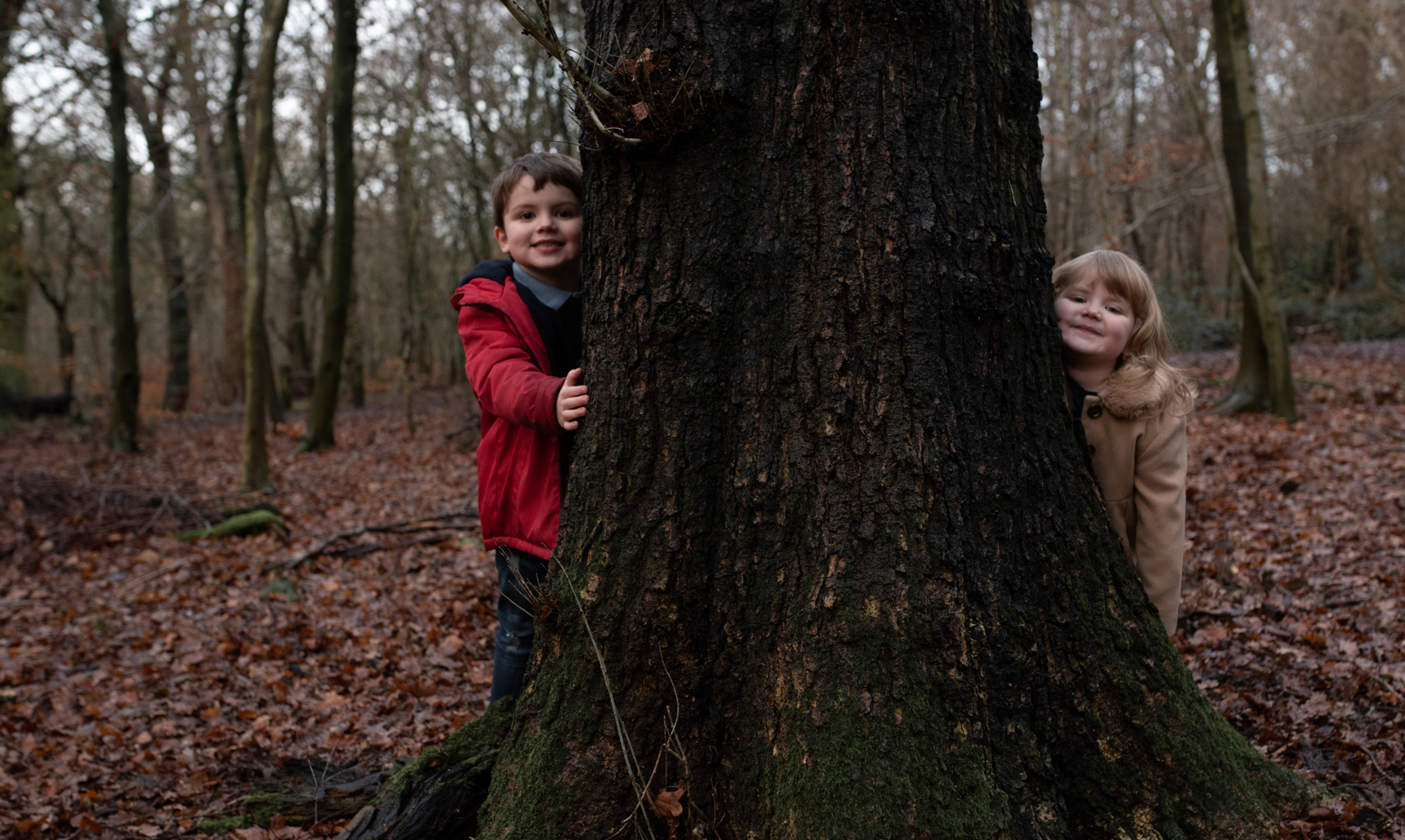 Two kids peeping around a tree in the woods