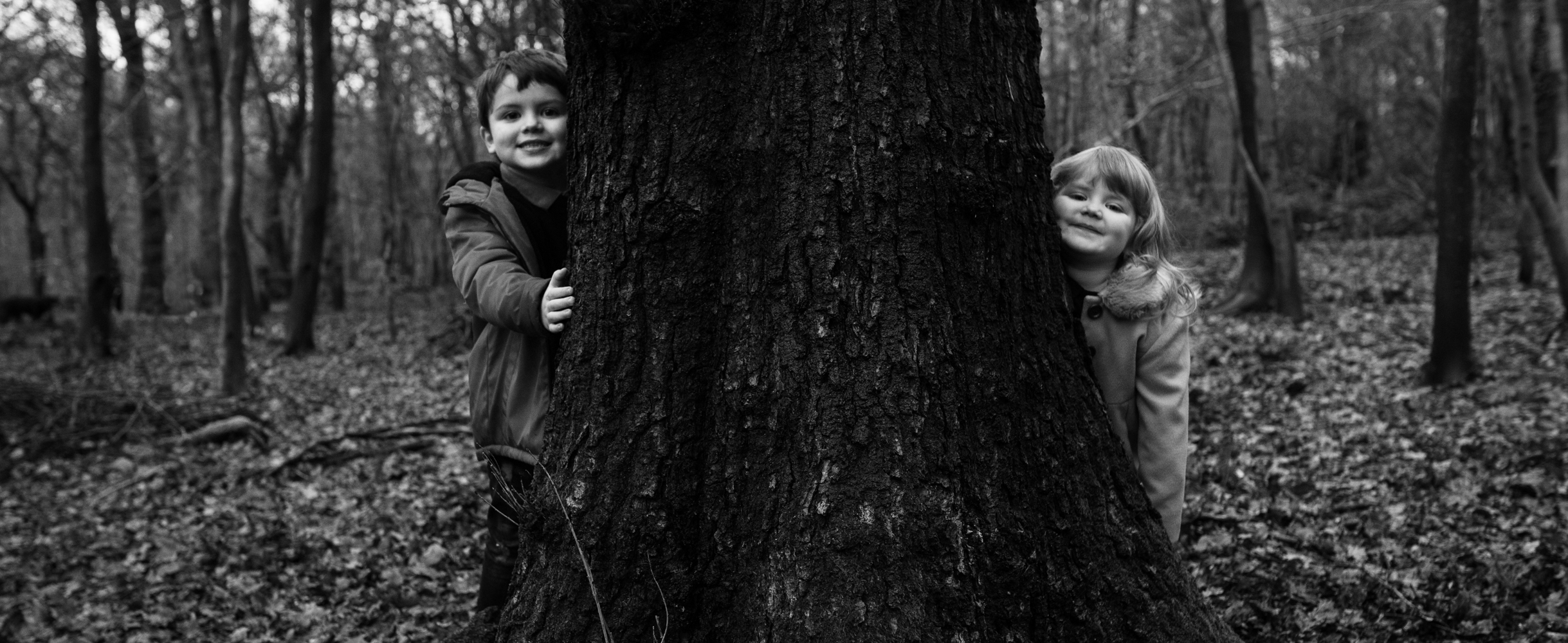 A black and white photograph of two kids peeping around a tree in the woods