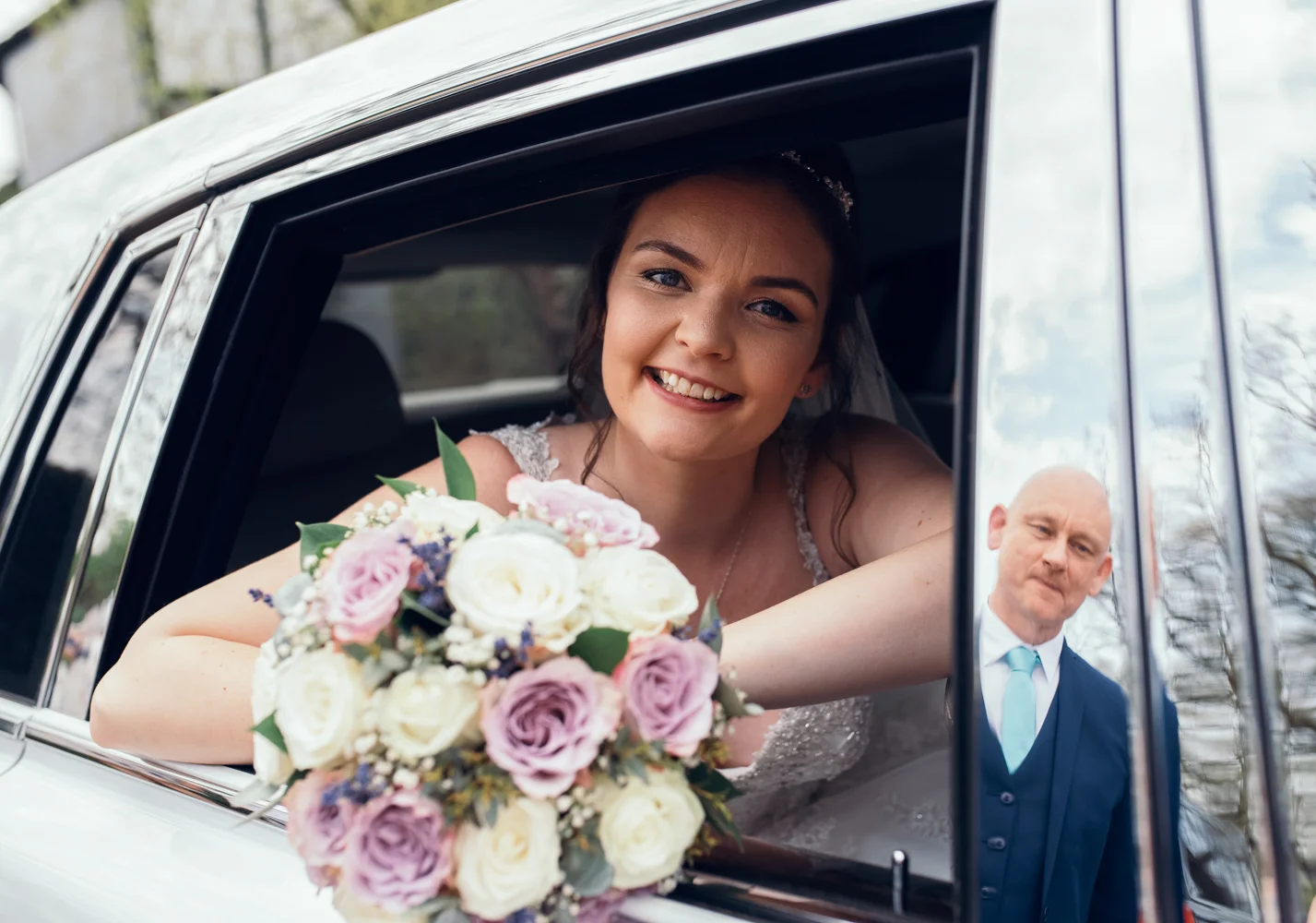 The bride sitting in the back of the car with her father reflected in a door panel
