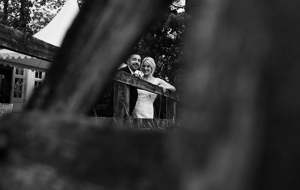 A black and white photo of the bride and groom taken through a small gap in the five bar wooden gate