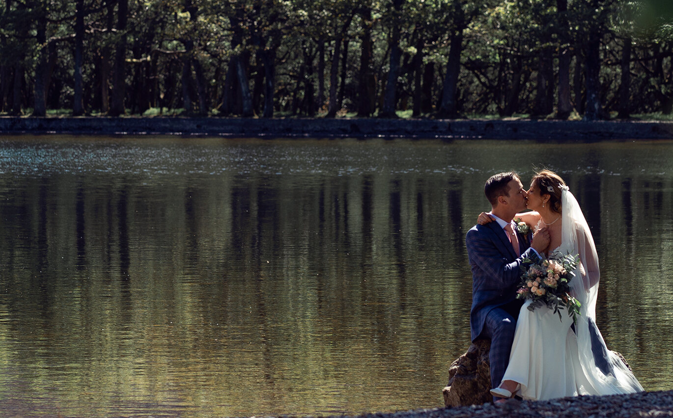 A bride and groom sitting on the lake shoreline kissing