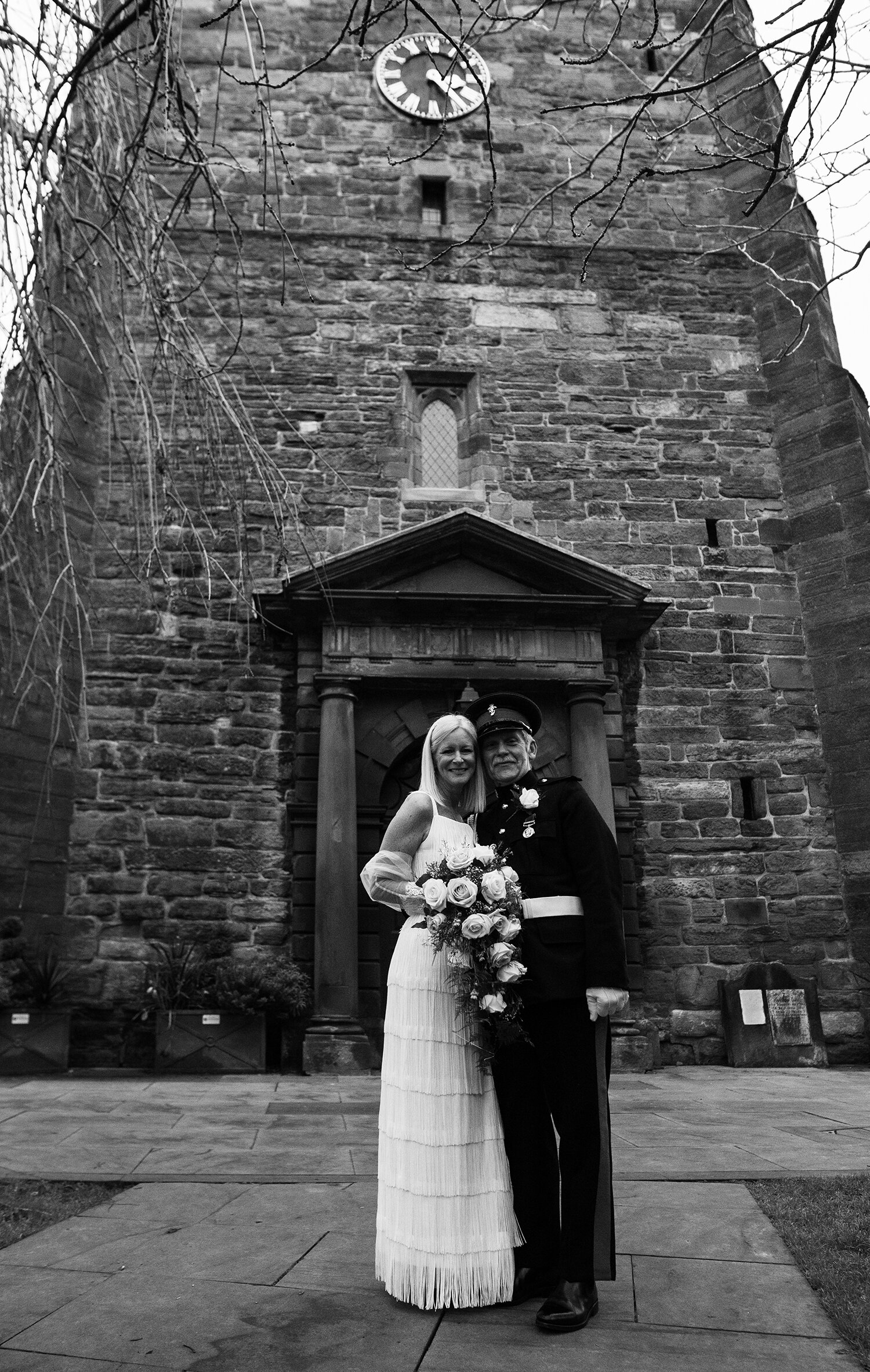 A black and white photo of the bride and groom standing in front of the church clock tower