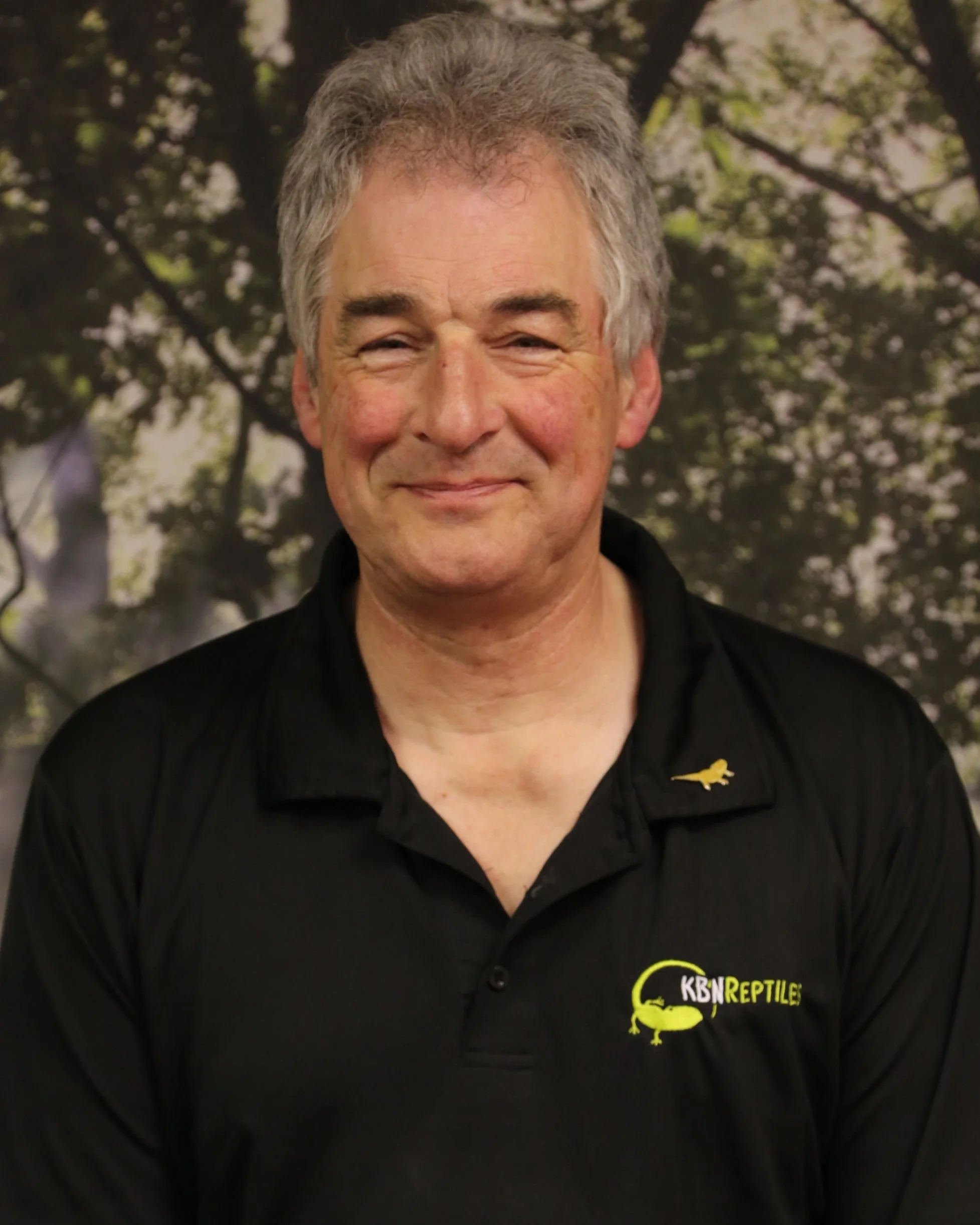 A middle-aged man with gray hair smiling, wearing a black polo shirt with a logo that reads 'KBN Reptiles' in yellow, with a yellow lizard graphic, standing outdoors in front of trees.