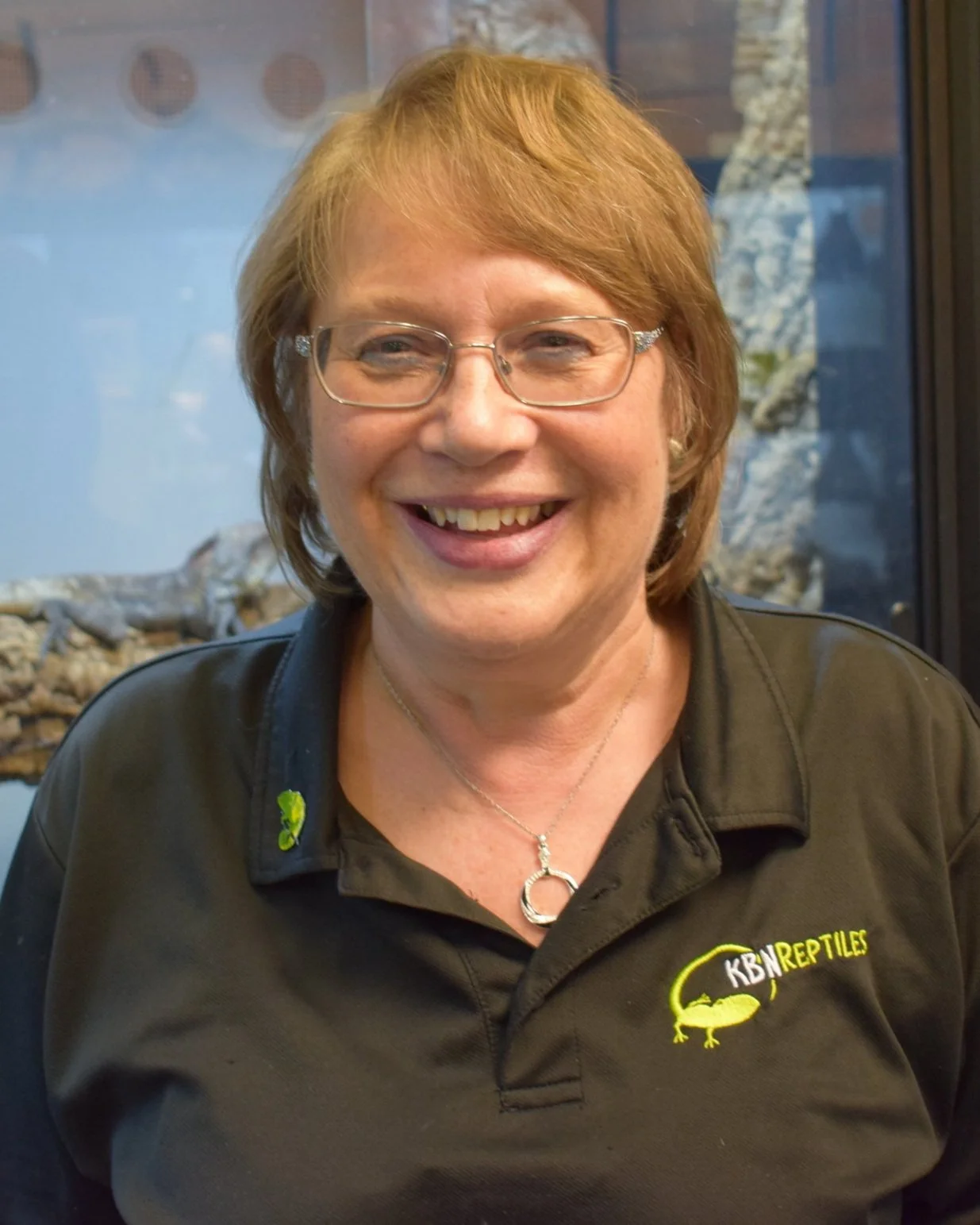A smiling middle-aged woman with short red hair and glasses wearing a black polo shirt with a 'KB Reptiles' logo, standing in front of a glass enclosure with reptiles inside.