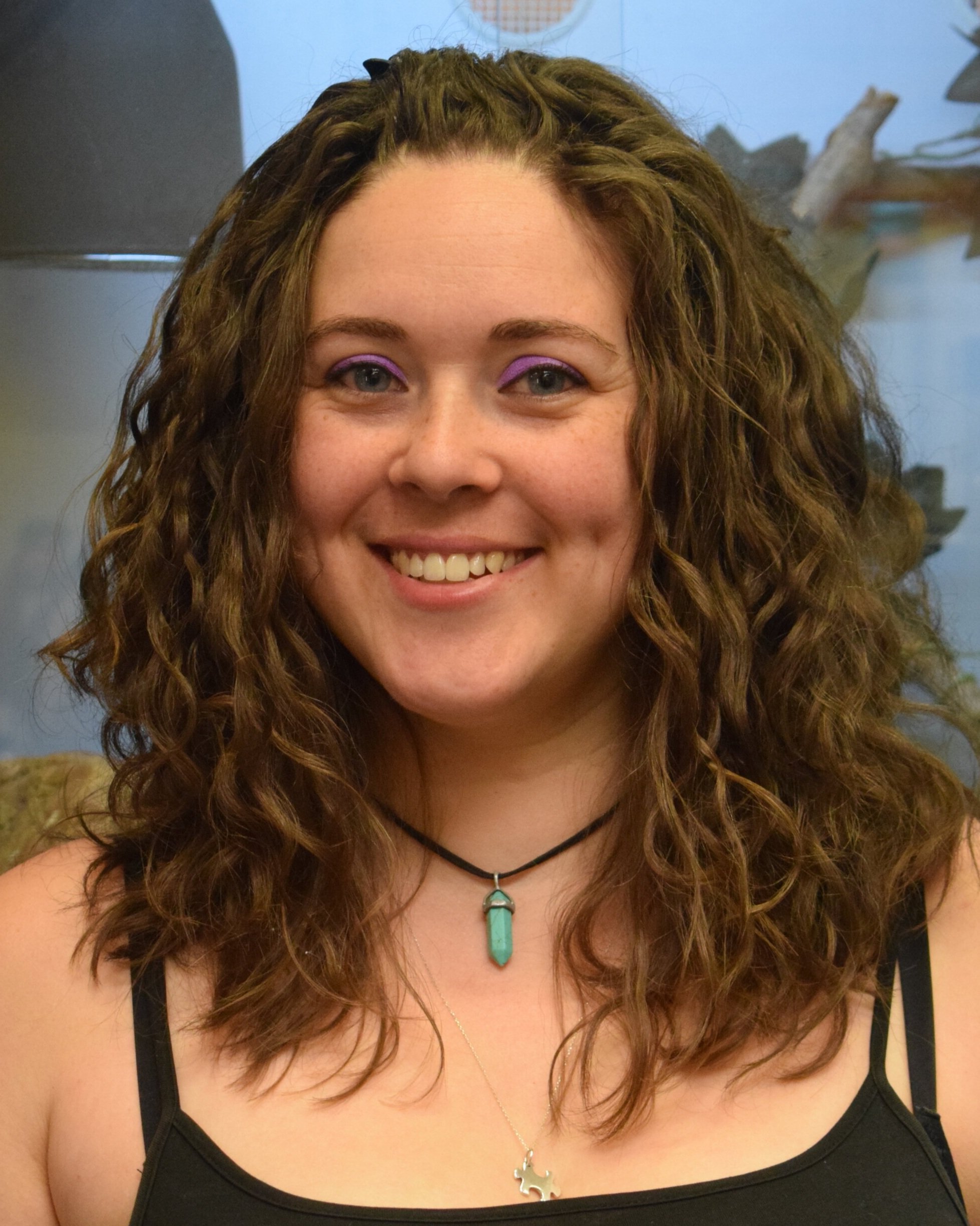 A smiling woman with curly brown hair, wearing purple eyeshadow, a black top with thin straps, a turquoise necklace with a pendant, and a gold chain. The background shows a blue sky, some greenery, and part of a building or garage.