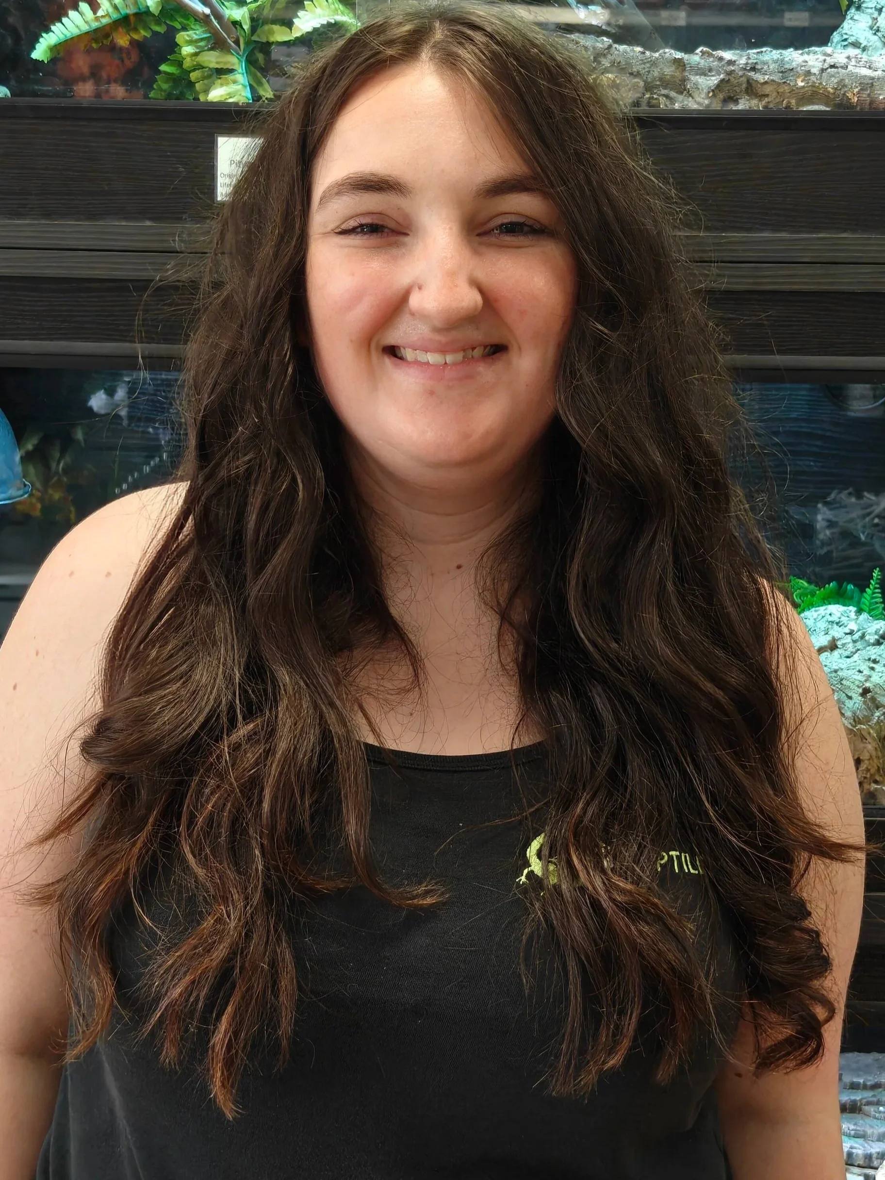 A woman with long, wavy brown hair and light makeup smiling. She is wearing a black shirt with green lettering and is sitting in front of an aquarium with green plants and rocks.