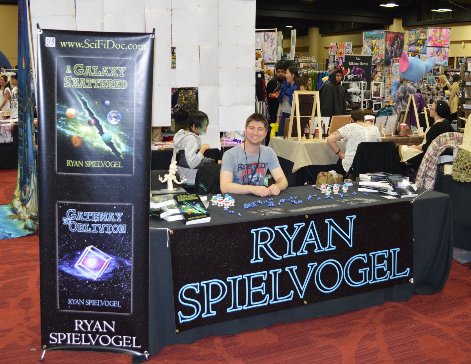 Me at my booth at Kraken Con with my assorted books and give-aways