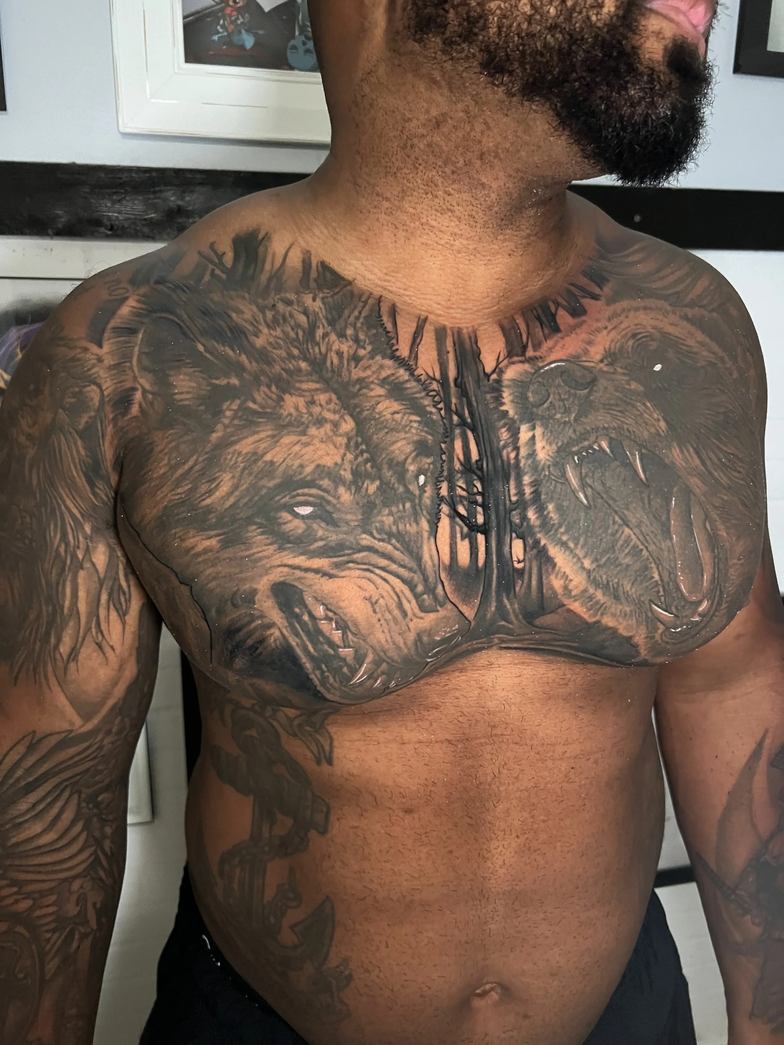 Tattooed man with a beard showing a large chest tattoo of two roaring wolves facing each other with a chain underneath them.