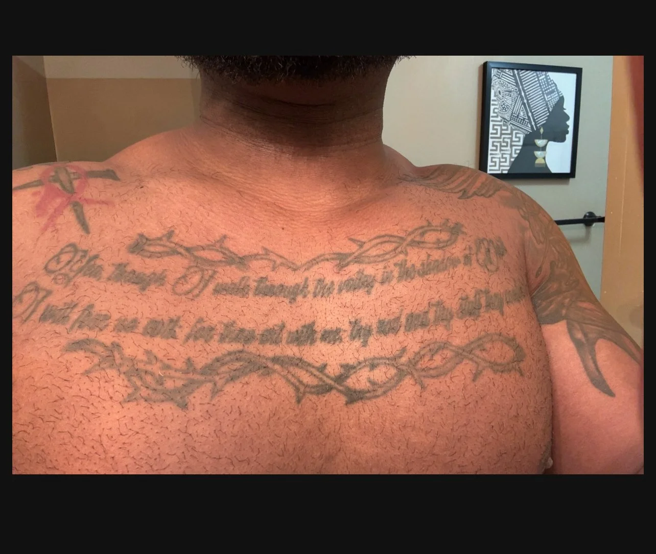 Close-up of a person's chest and shoulders with multiple tattoos, including a barbed wire design and script text, in a bathroom with framed artwork in the background.