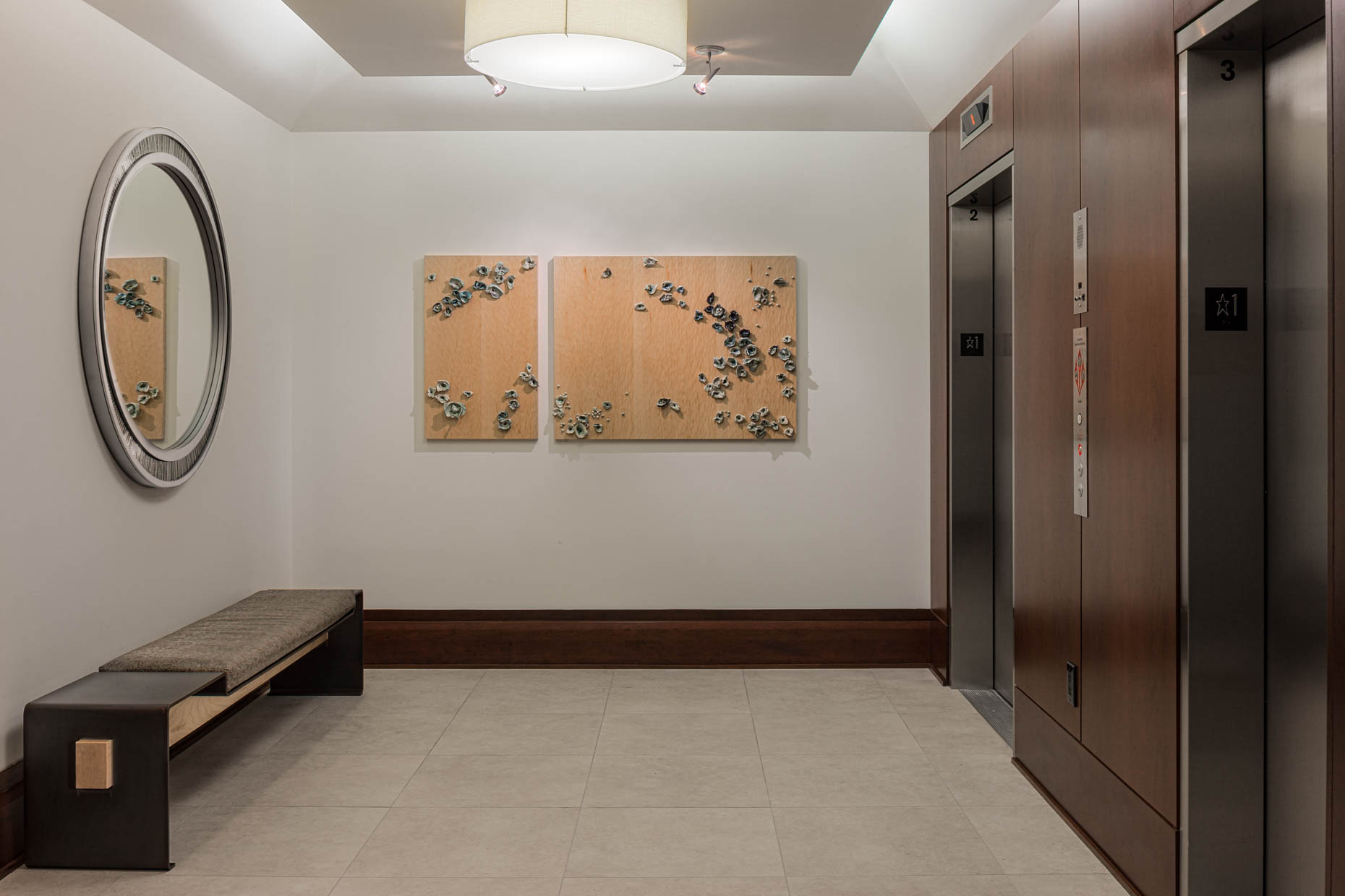condo lobby showing bright white tile floor, up-lighting, a maple, steel and leather bench, oval steel and porcelain mosaic mirror and wood wall panels