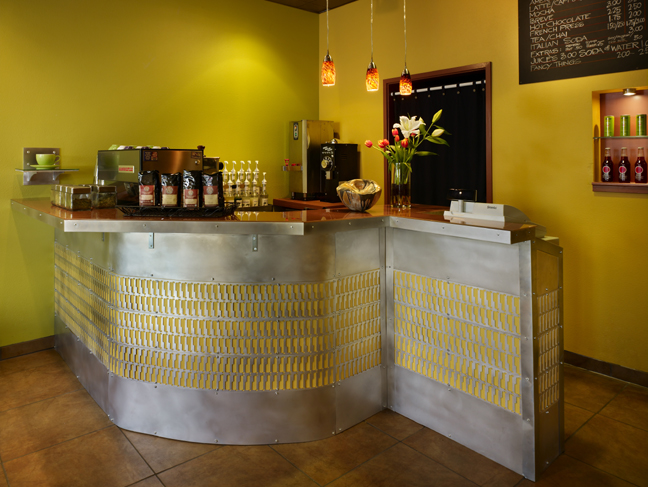 Coffee counter in cafe with steel inlay, back-painted glass tops, a product niche beyond showing colorful bottles and cans, a handmade bowl, a vase of flowers and pendant lighting