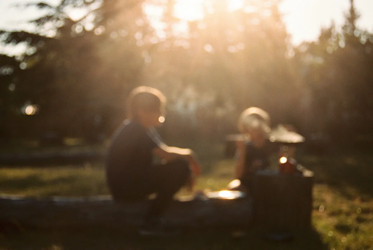 two boys sitting in sun flare in campsite cooking 