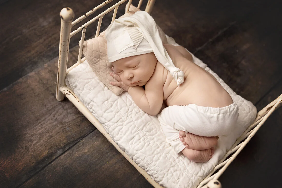 newborn with cream sleepy hat on iron bed prop