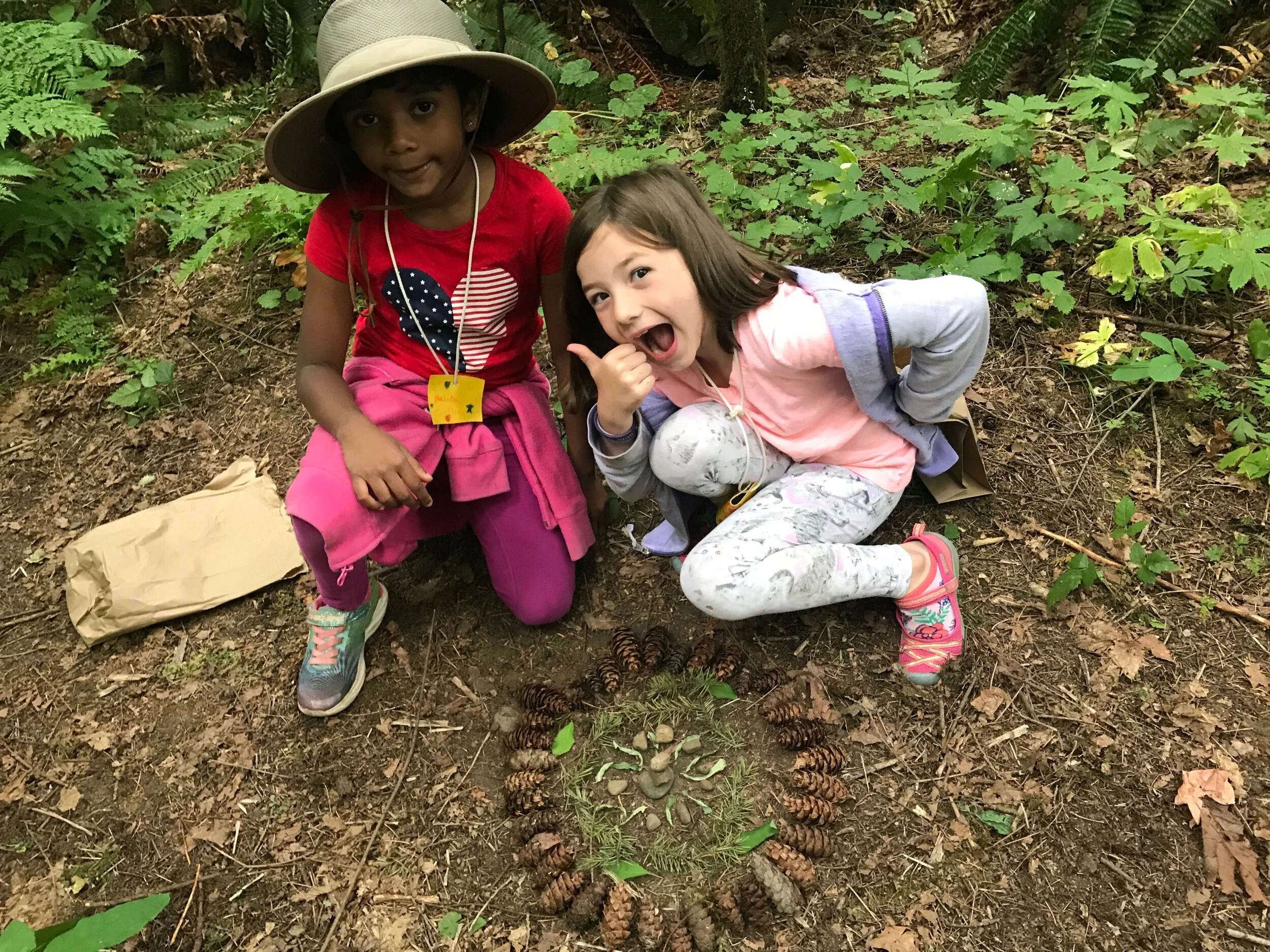  Two students crouch on the forest floor by their pinecone art 